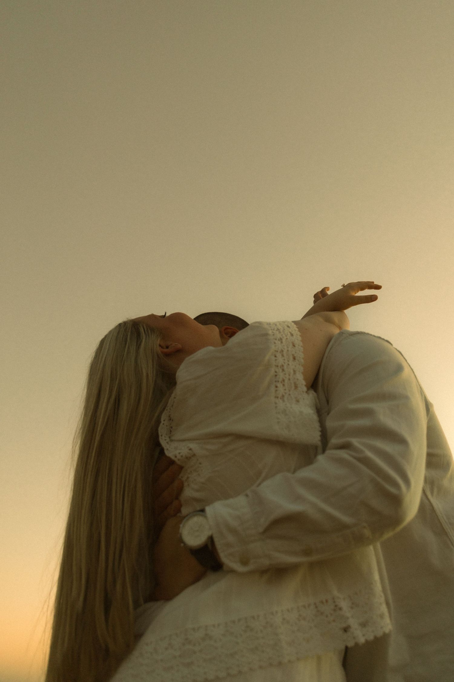 Couple in white clothing sharing a romantic kiss at golden hour sunset, woman with long blonde hair.
