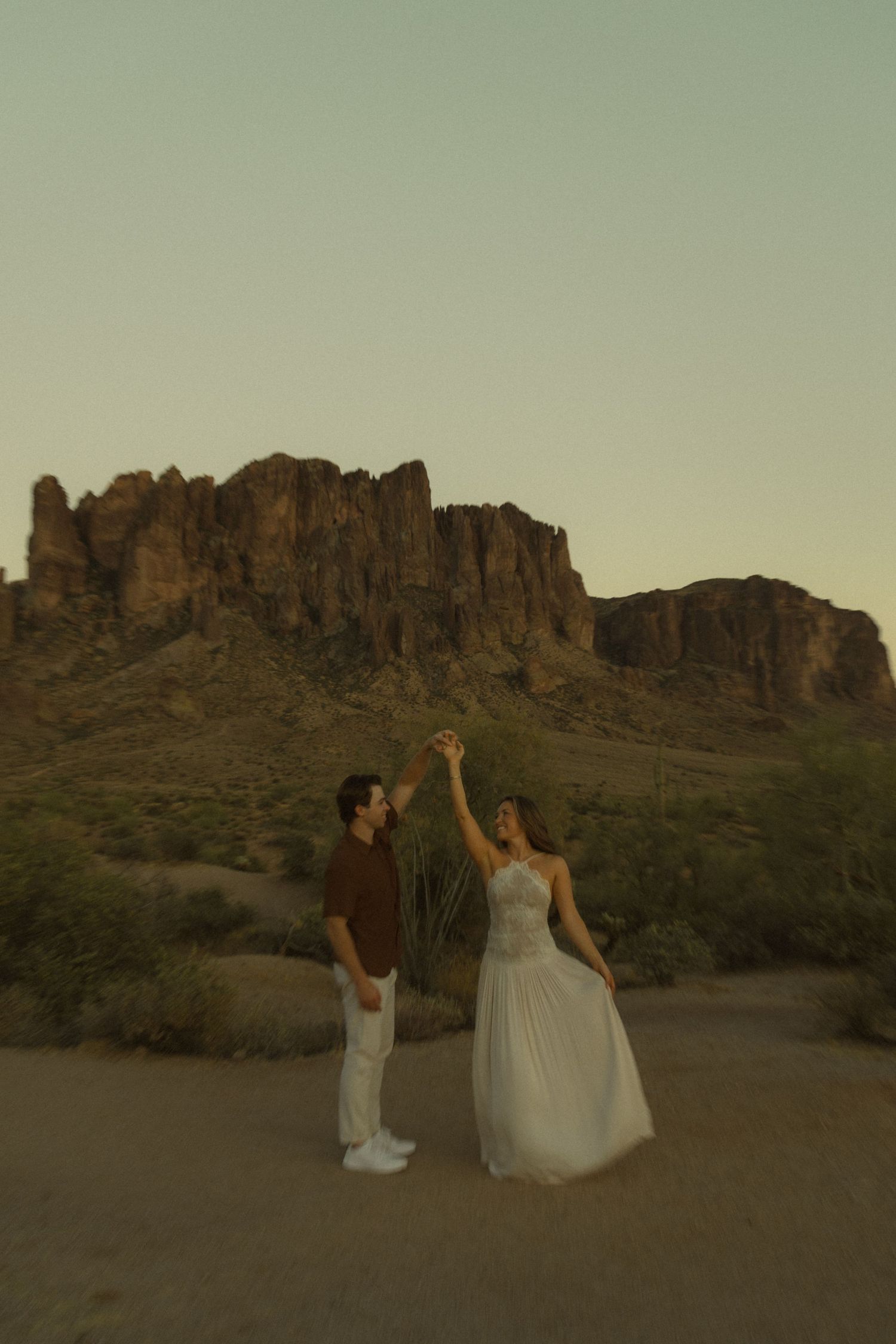 Couple dancing together in white and brown outfits with man spinning woman in Arizona desert at sunset.