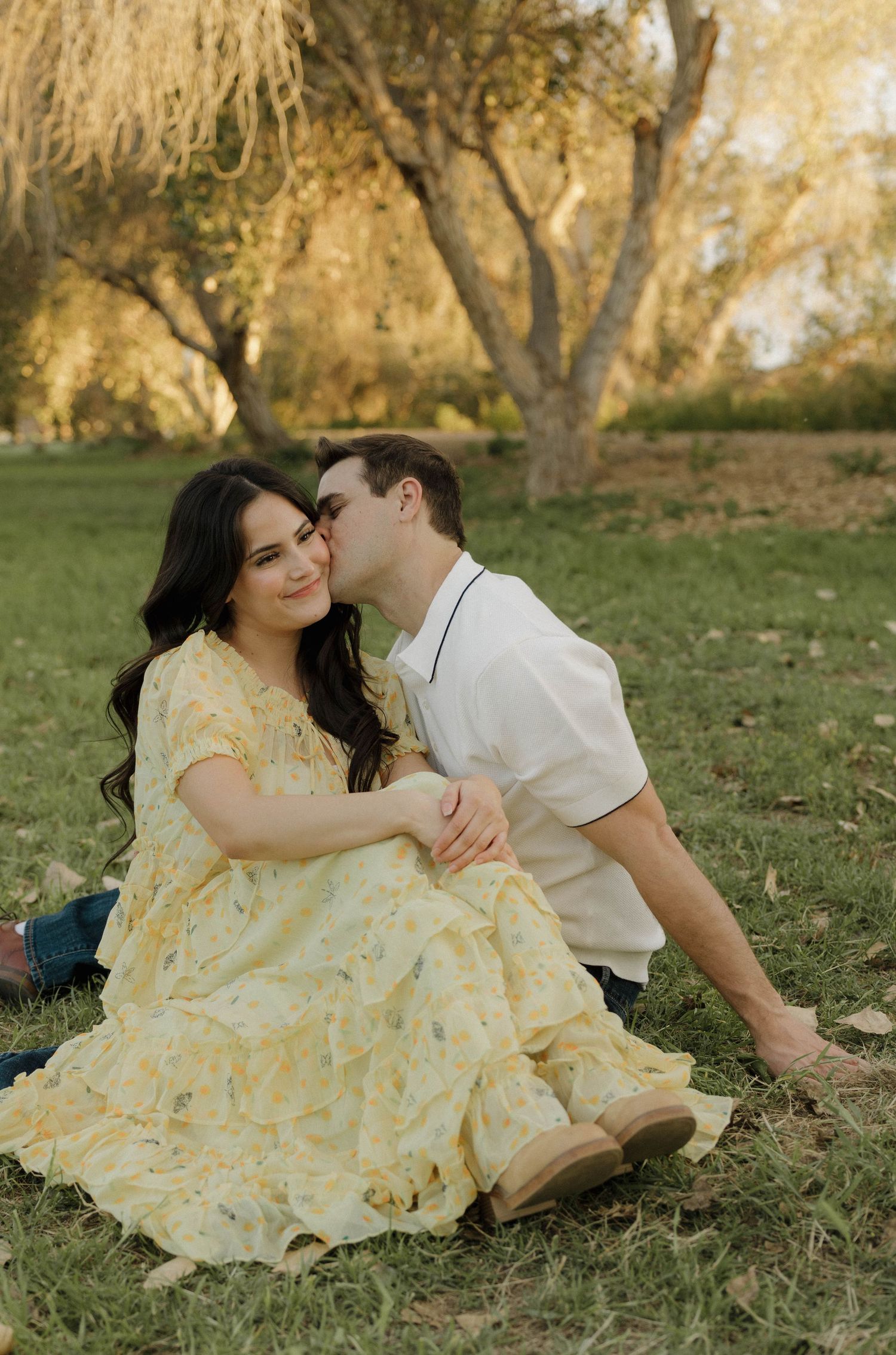 A romantic couple's photoshoot showing intimate moments on grass under golden sunlit trees.