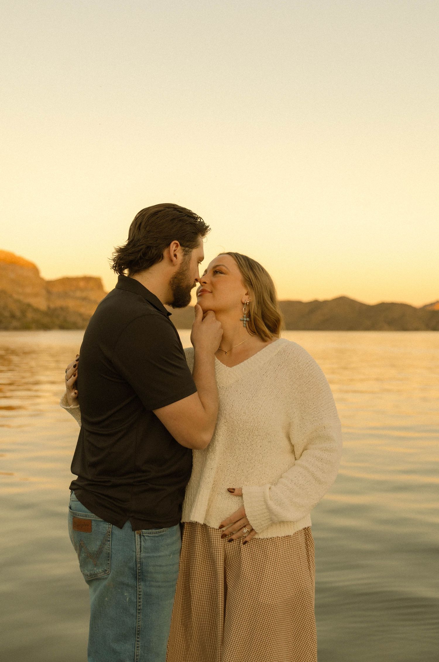 A romantic couple shares a tender kiss at sunset by a serene lake with mountains in the background.
