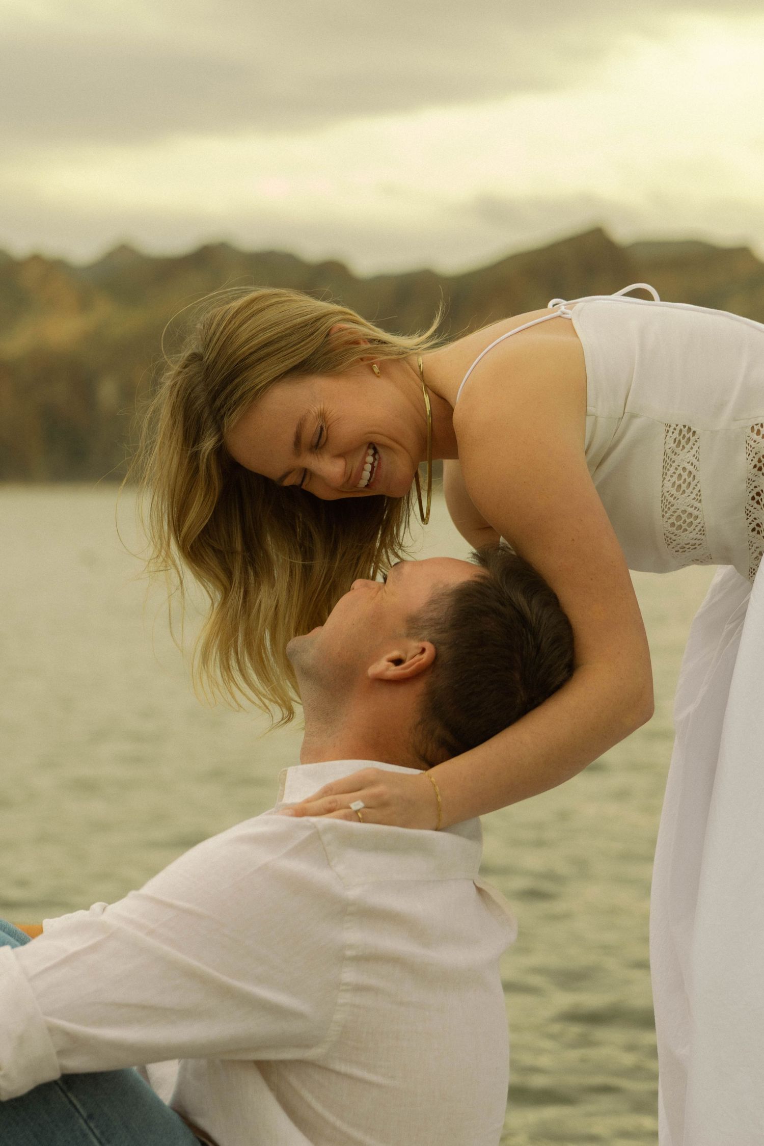 A romantic couple embraces joyfully by a lake with mountains in the background during golden hour.