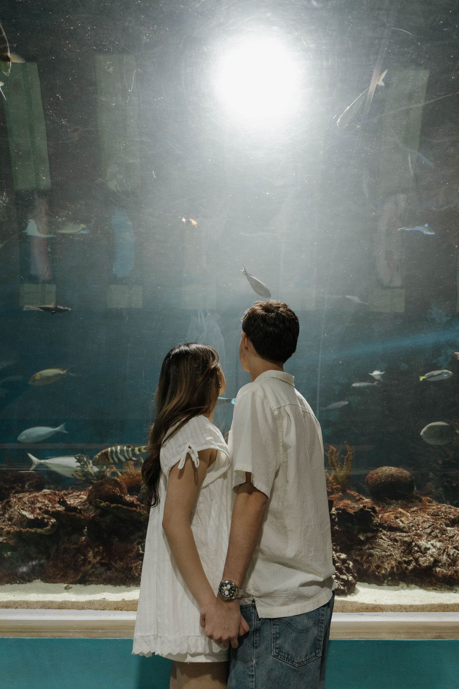 A couple in light colored outfits views fish swimming in a dimly lit aquarium tank.