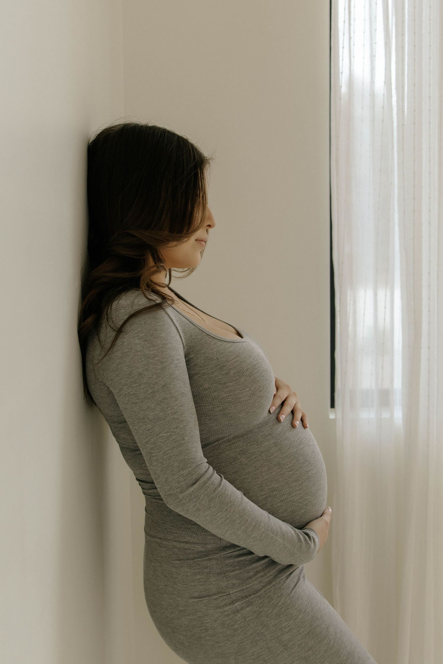 A pregnant woman in a grey dress stands in profile by a window with sheer curtains.