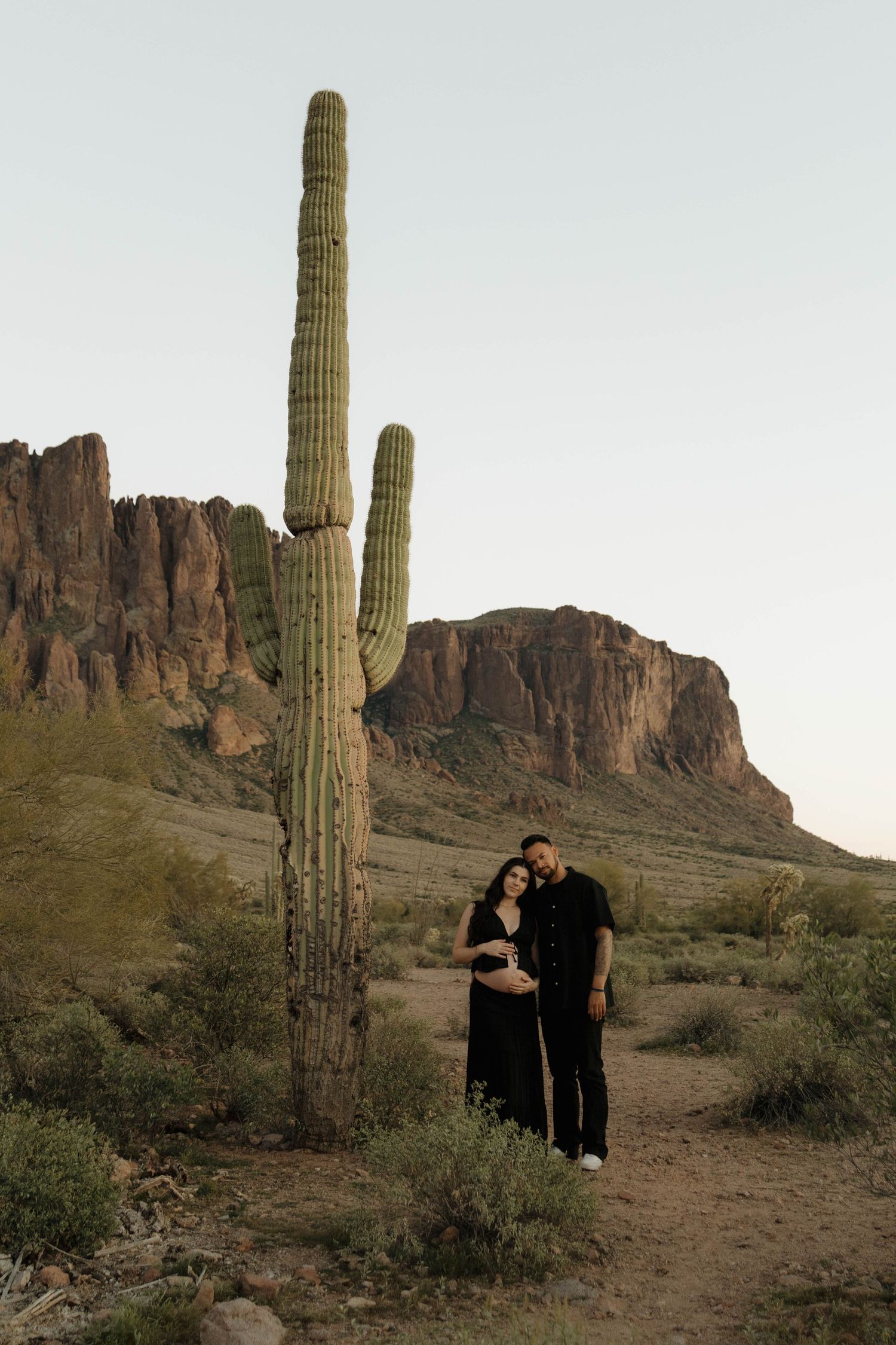 Couple embracing near tall saguaro cactus with desert cliffs in background during golden hour.