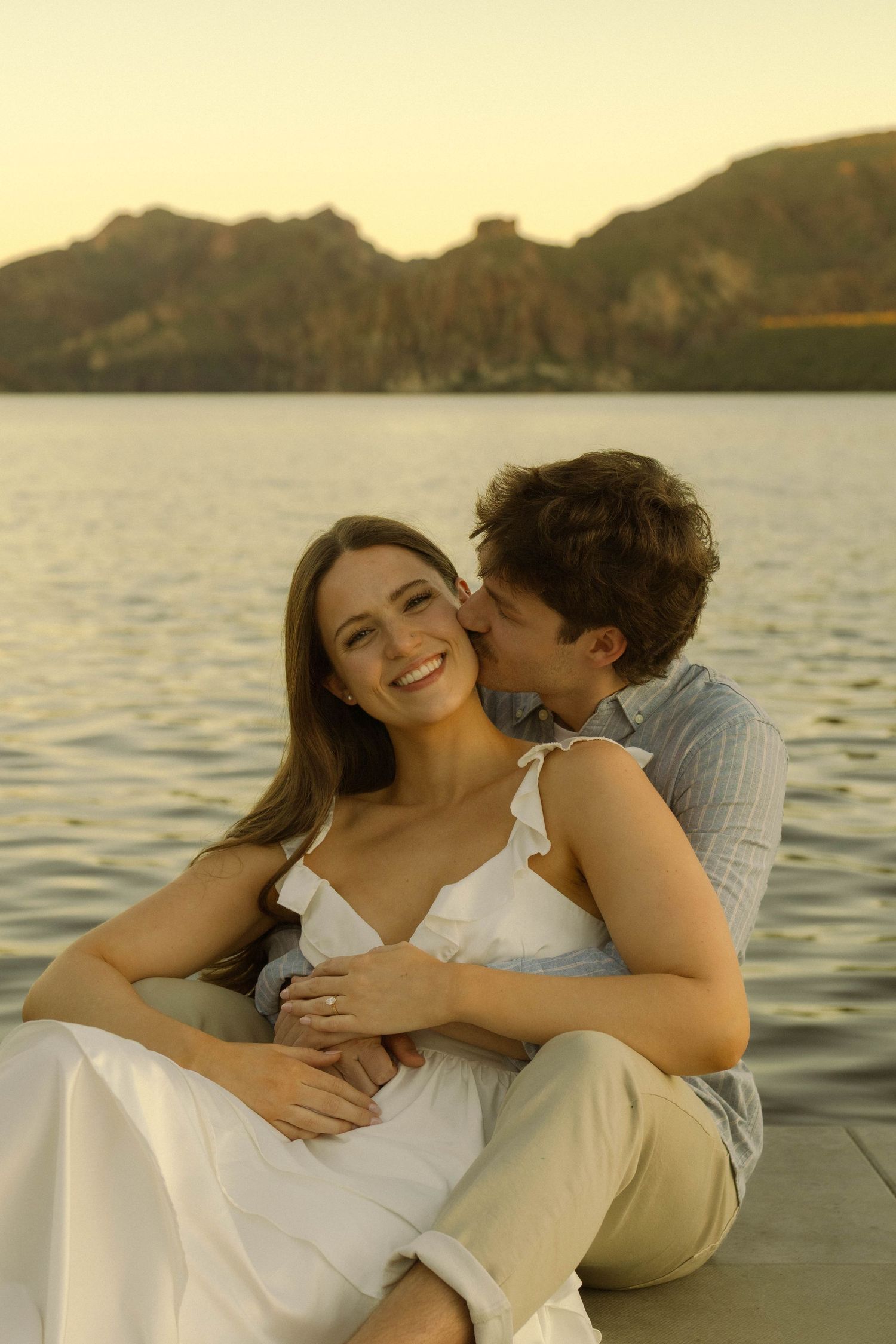 A romantic couple shares an intimate moment by a lake with mountains in the background during golden hour.