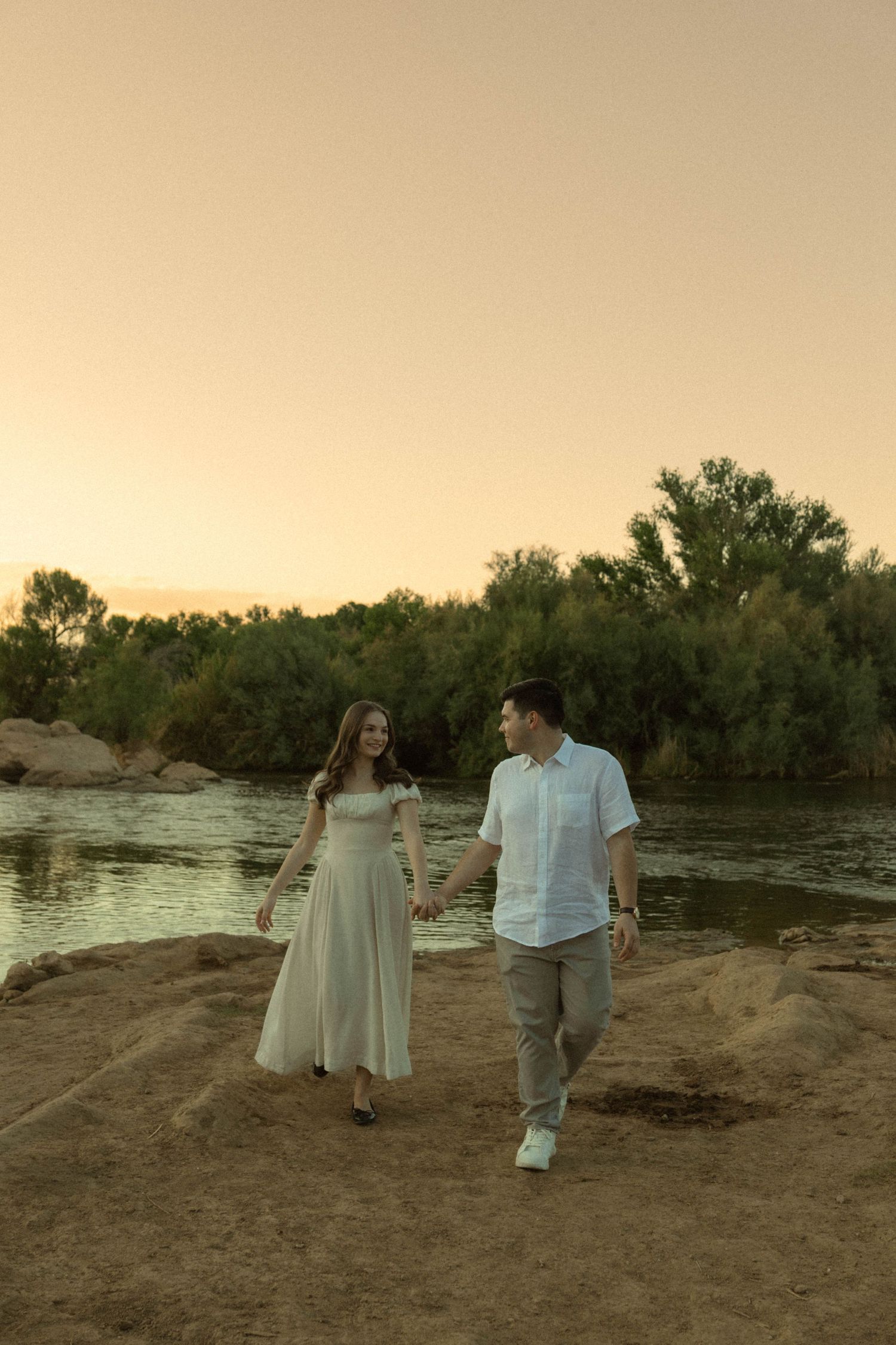 Couple holding hands walking along a rocky riverbank at golden sunset with lush green trees in background.