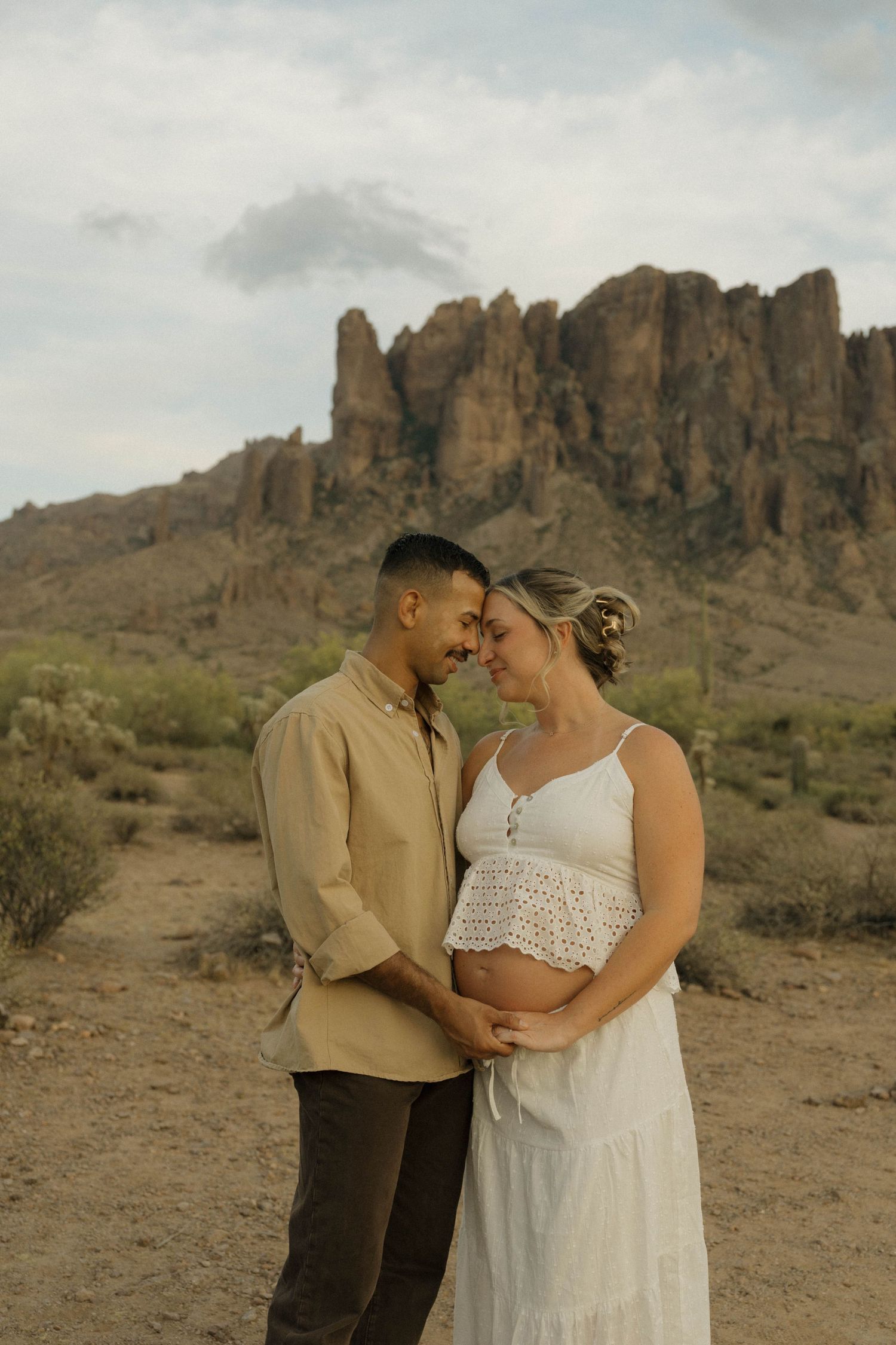 Expecting couple touch foreheads tenderly in Sonoran Desert landscape.