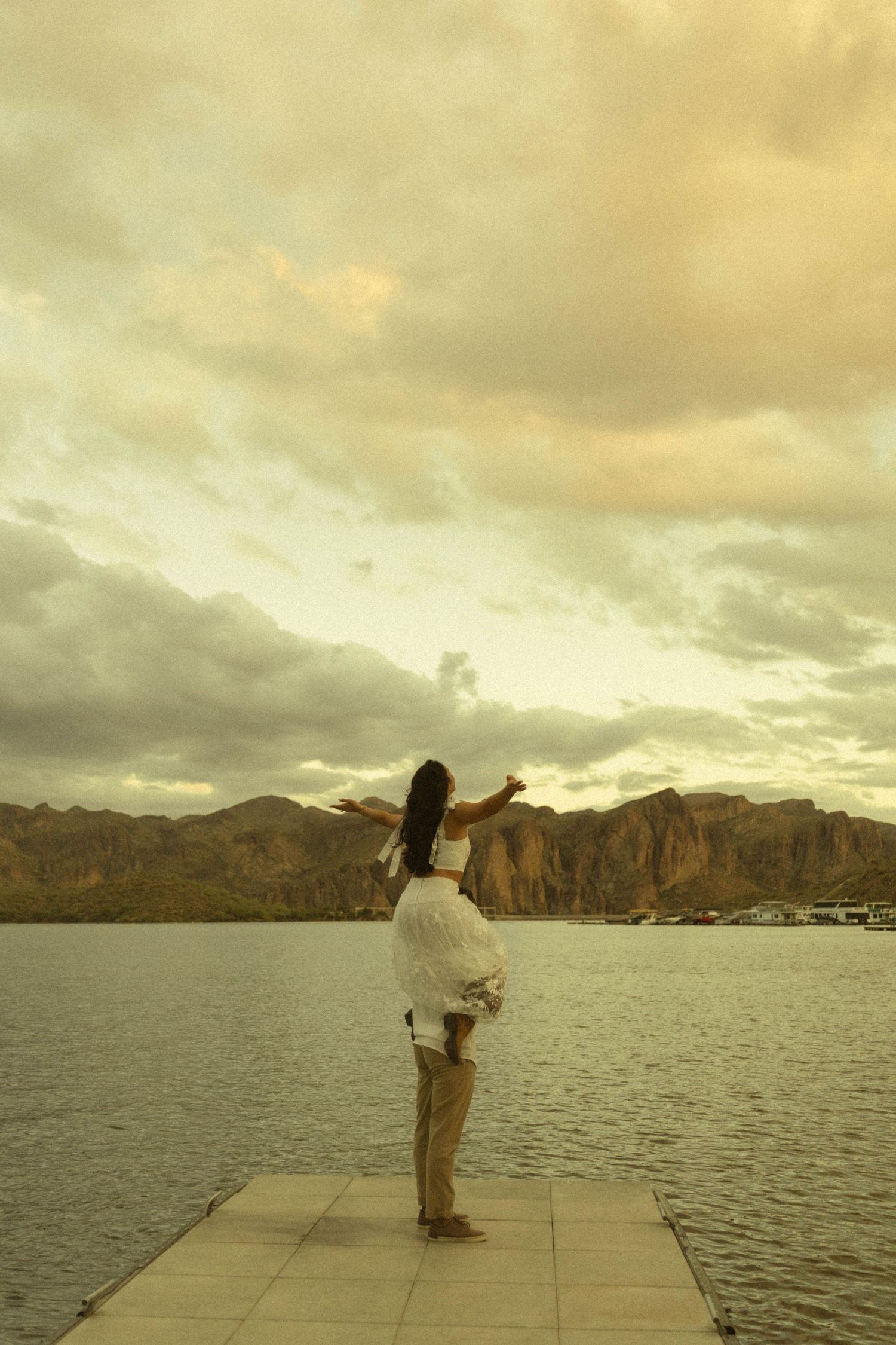 Woman in white dress stands on dock with arms outstretched, facing mountain lake under dramatic cloudy sunset sky.