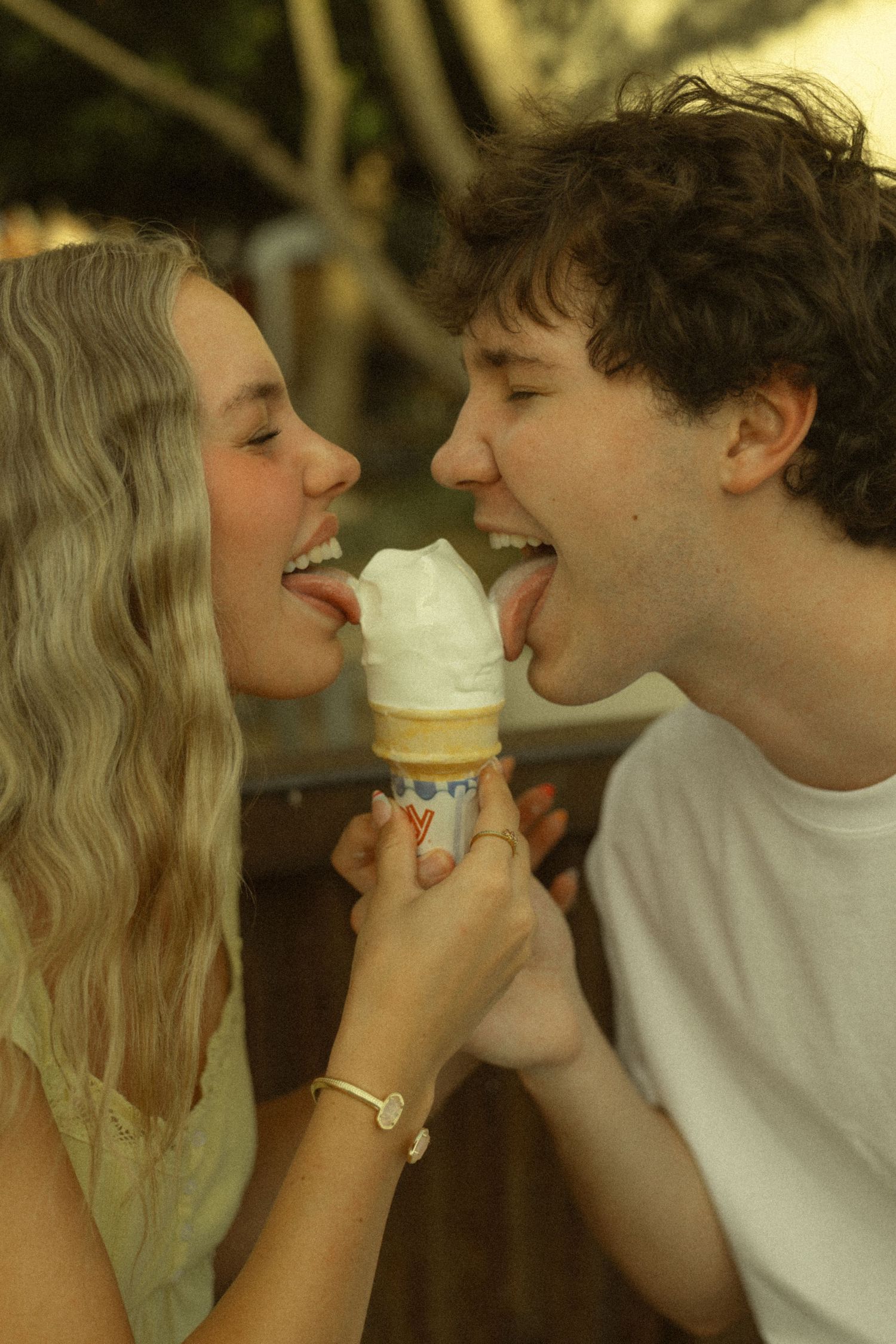 Young couple sharing a soft serve ice cream cone together, both smiling and licking it simultaneously outdoors.