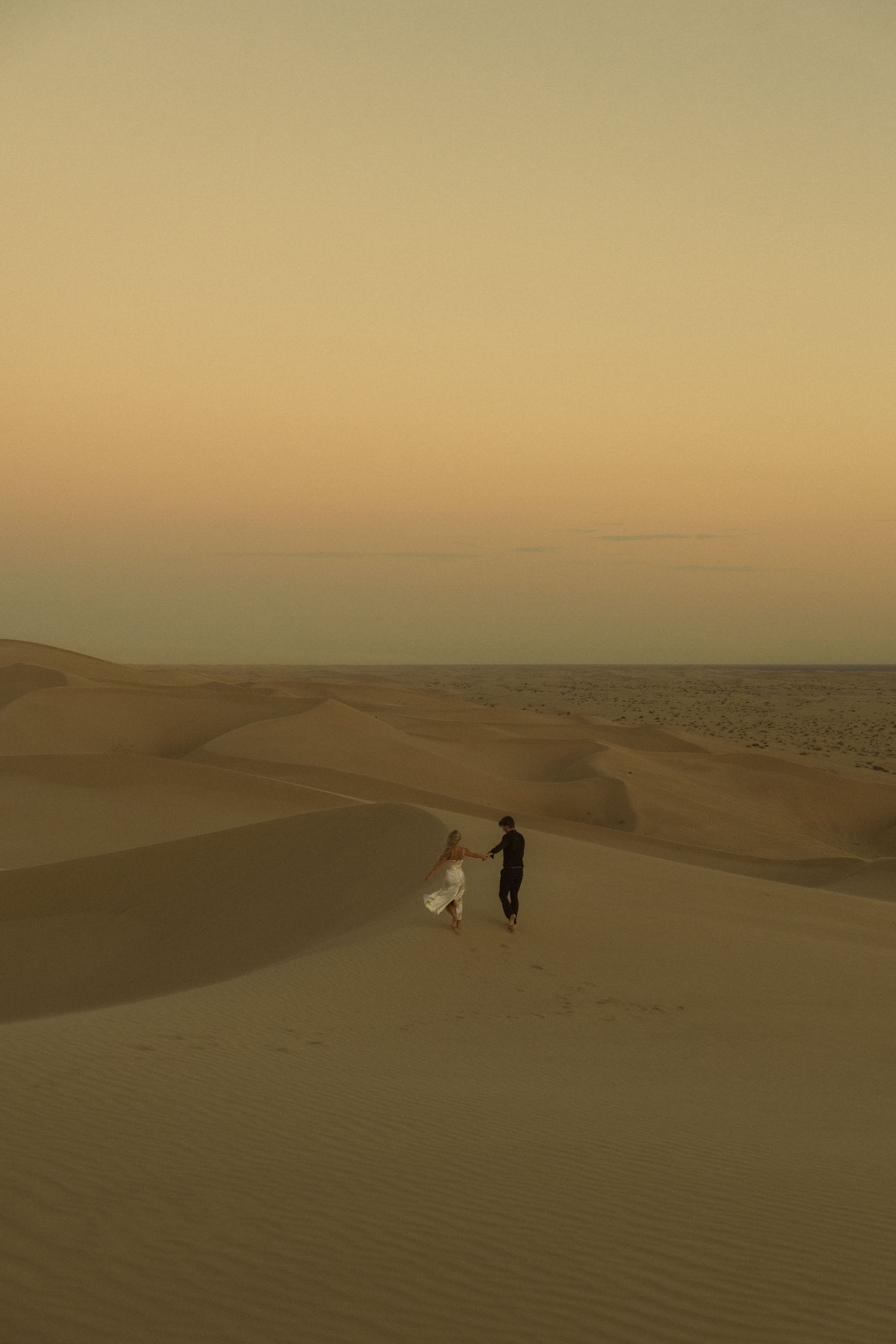 Couple holding hands walking across vast golden sand dunes at dusk in desert.