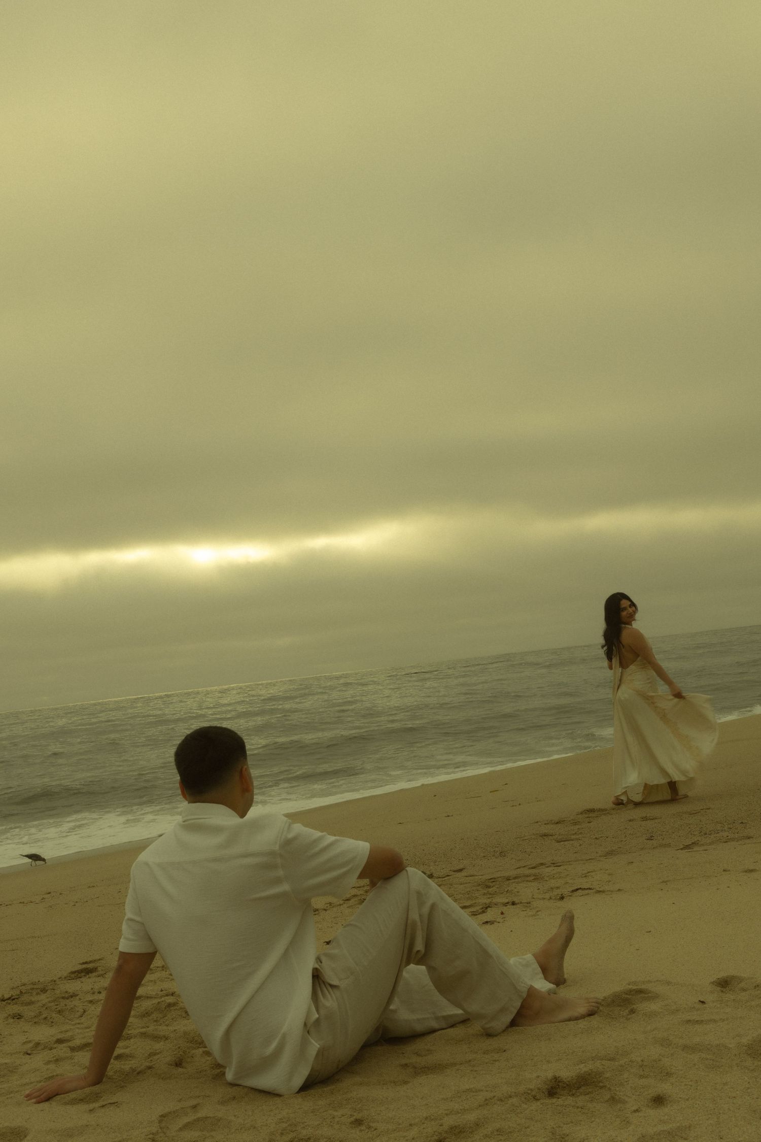 Couple in white outfits on a misty beach at sunset, man sitting on sand watching woman in flowing dress near the ocean.