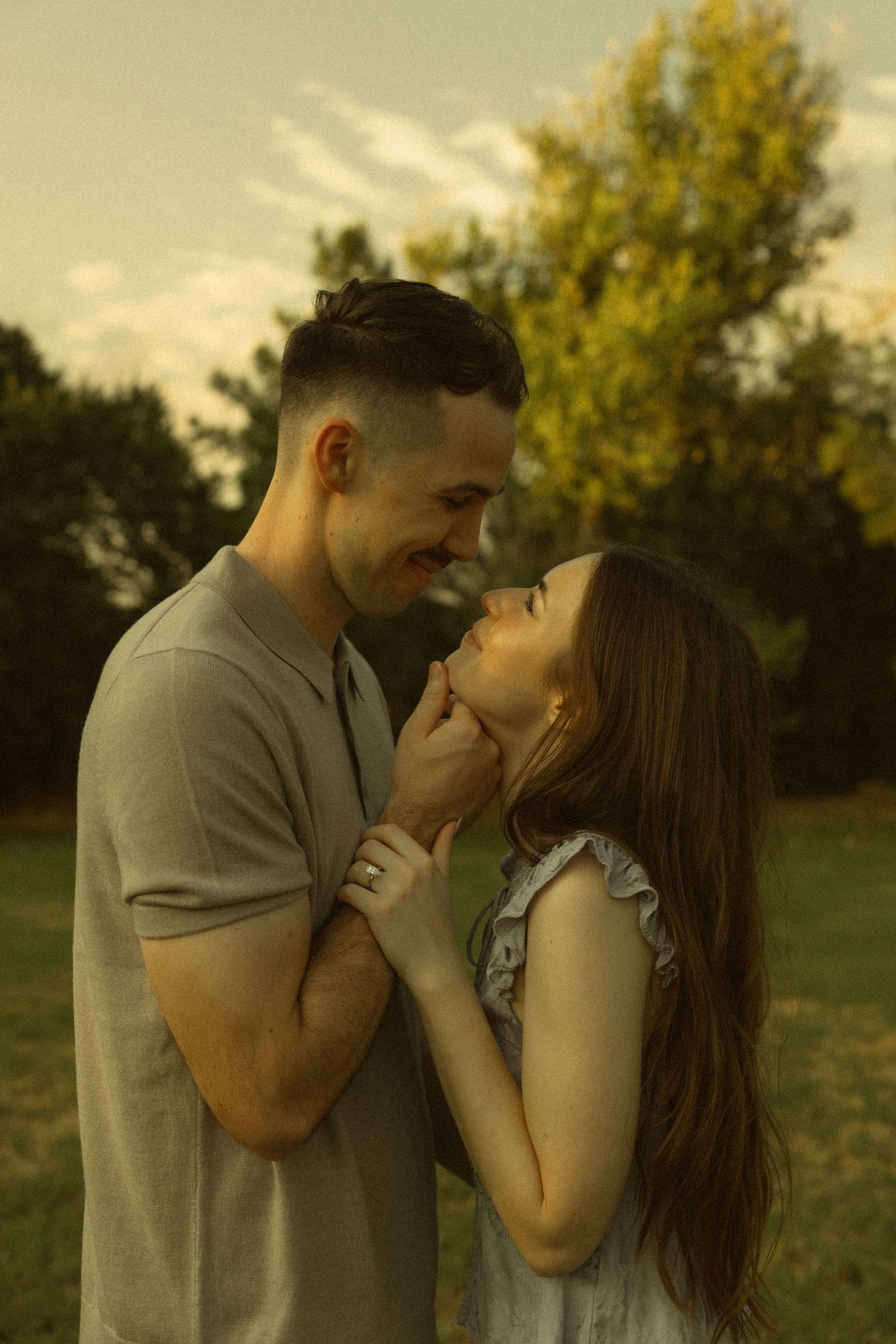Romantic couple holding hands and gazing at each other in a park with trees in background.