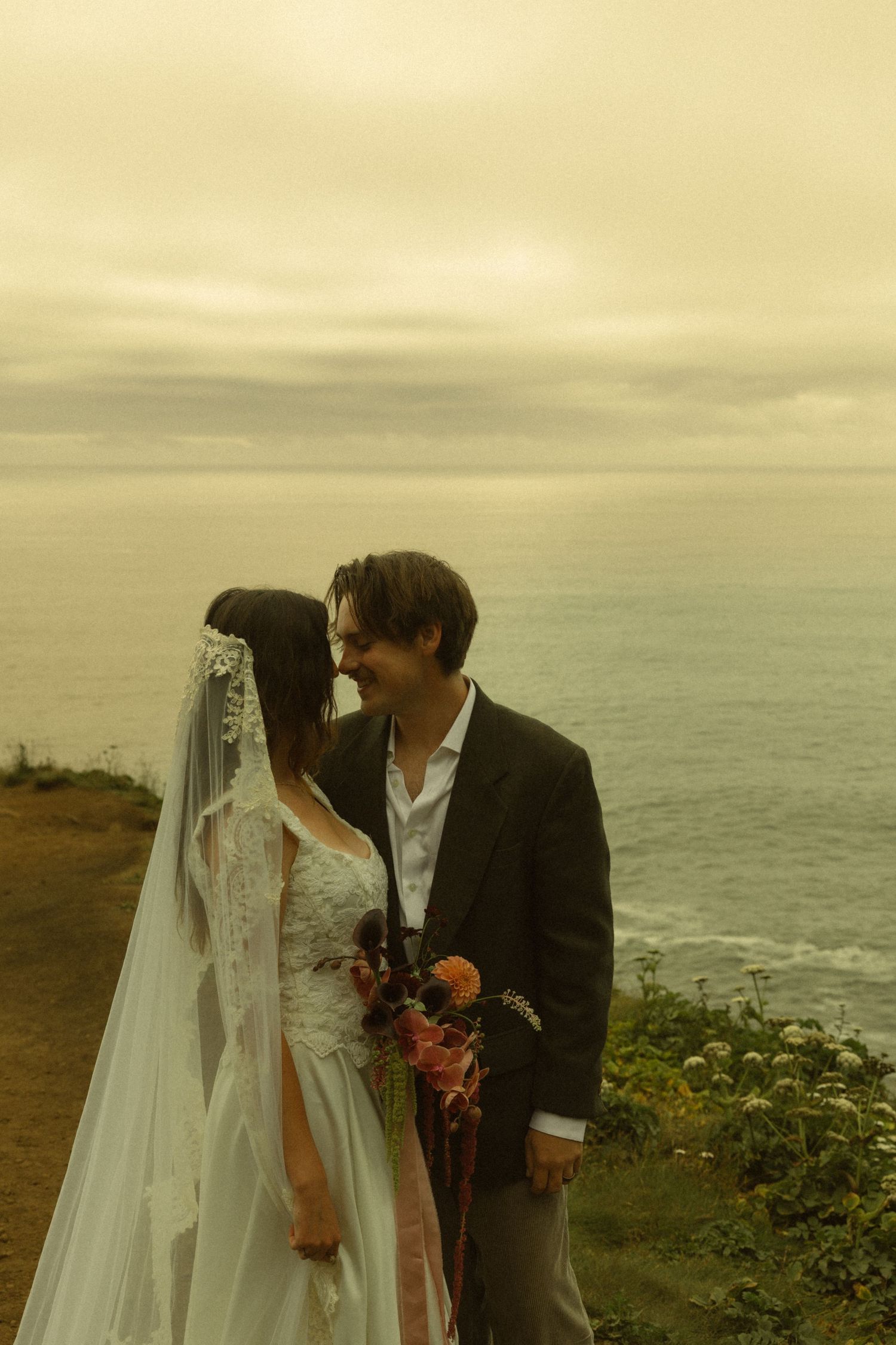 Bride in lace gown and veil holding bouquet, sharing intimate moment with groom by the ocean.