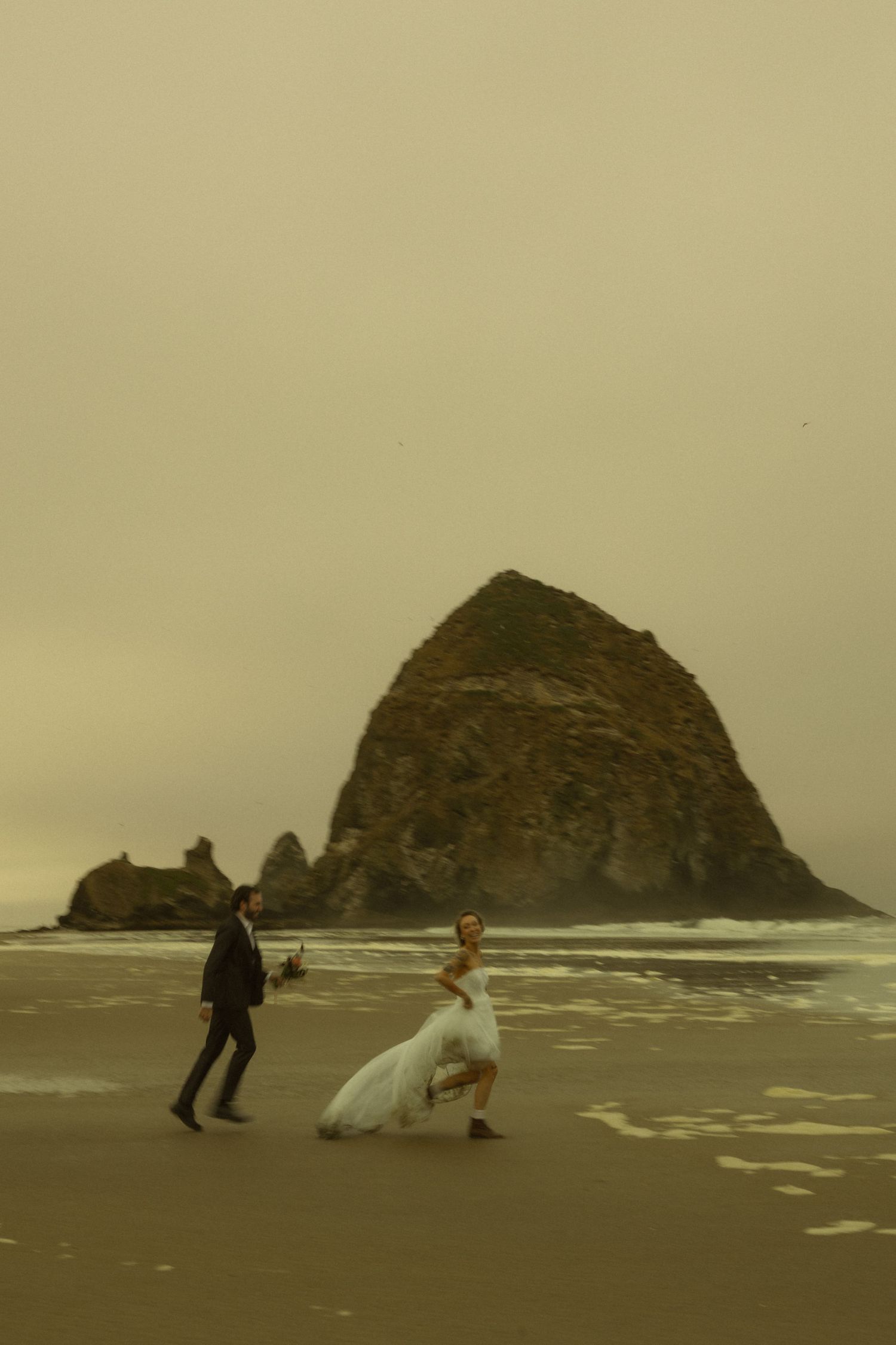 Bride in white gown and groom in dark suit walk along Cannon Beach with Haystack Rock in the misty background.