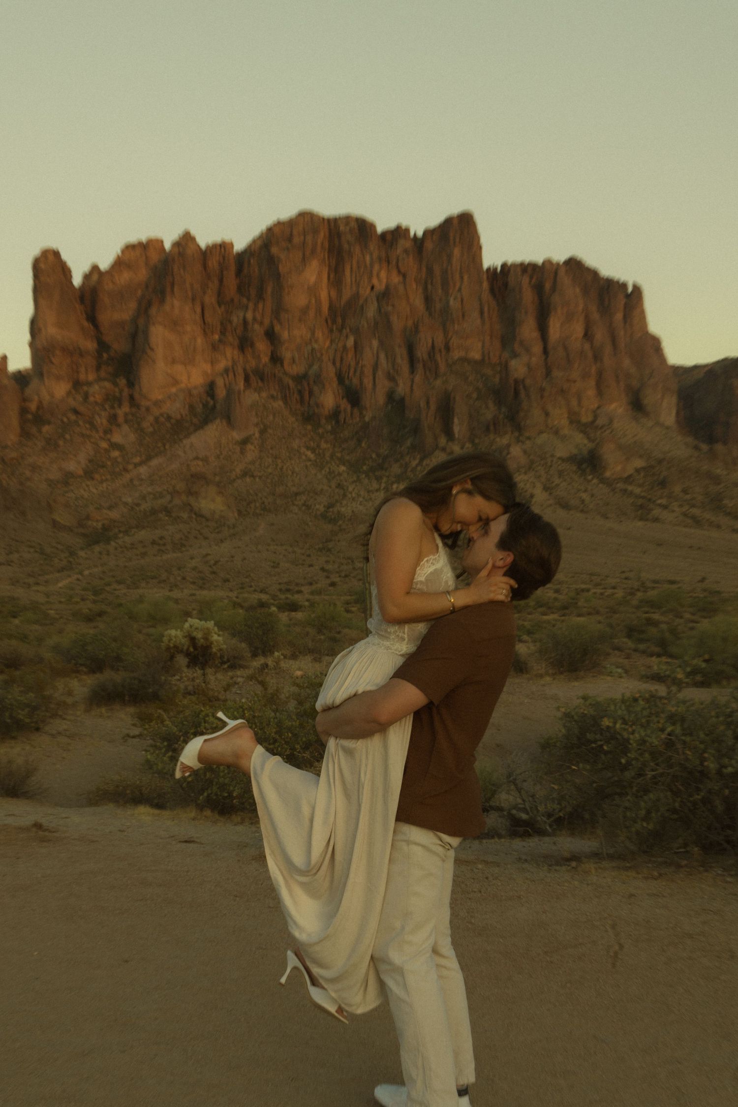 Couple sharing a romantic kiss at sunset in the Arizona desert with dramatic Superstition Mountains in the background.