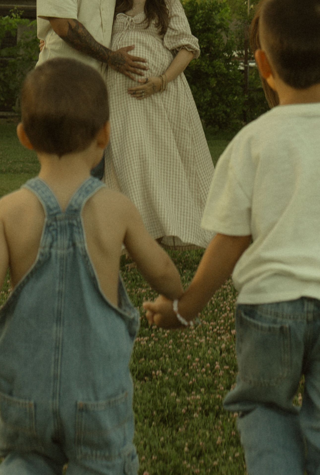 Two young boys in denim hold hands facing a pregnant woman in a floral field during a family maternity photo session.