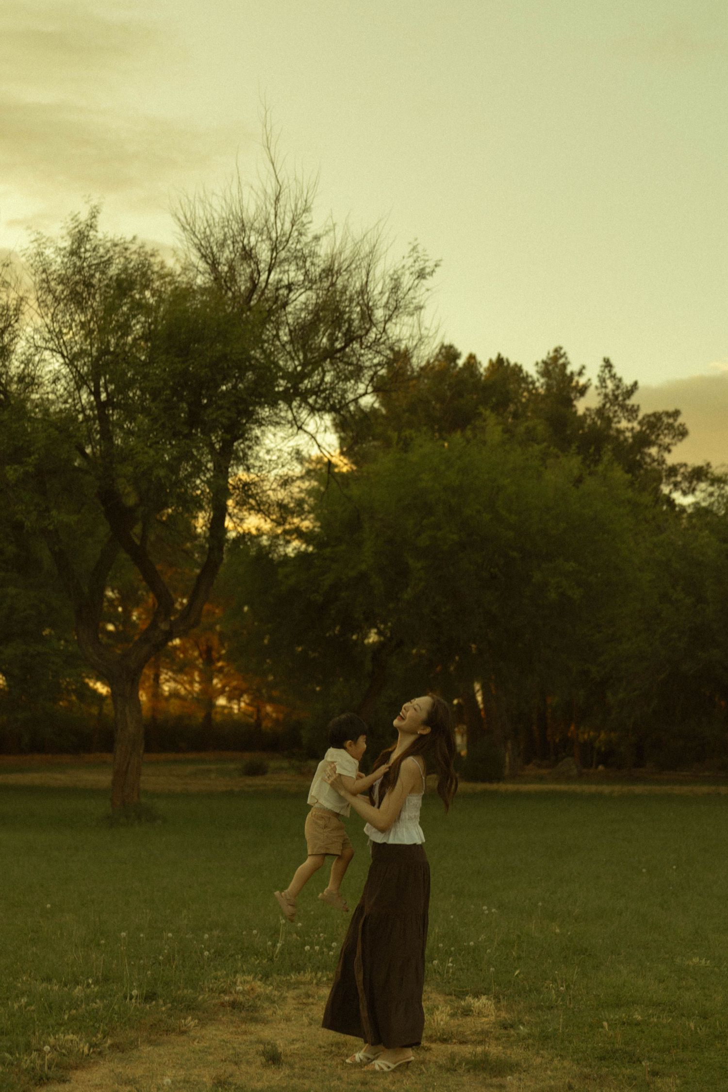 A mother lifts her baby up while standing in a park at sunset, looking up with joy.