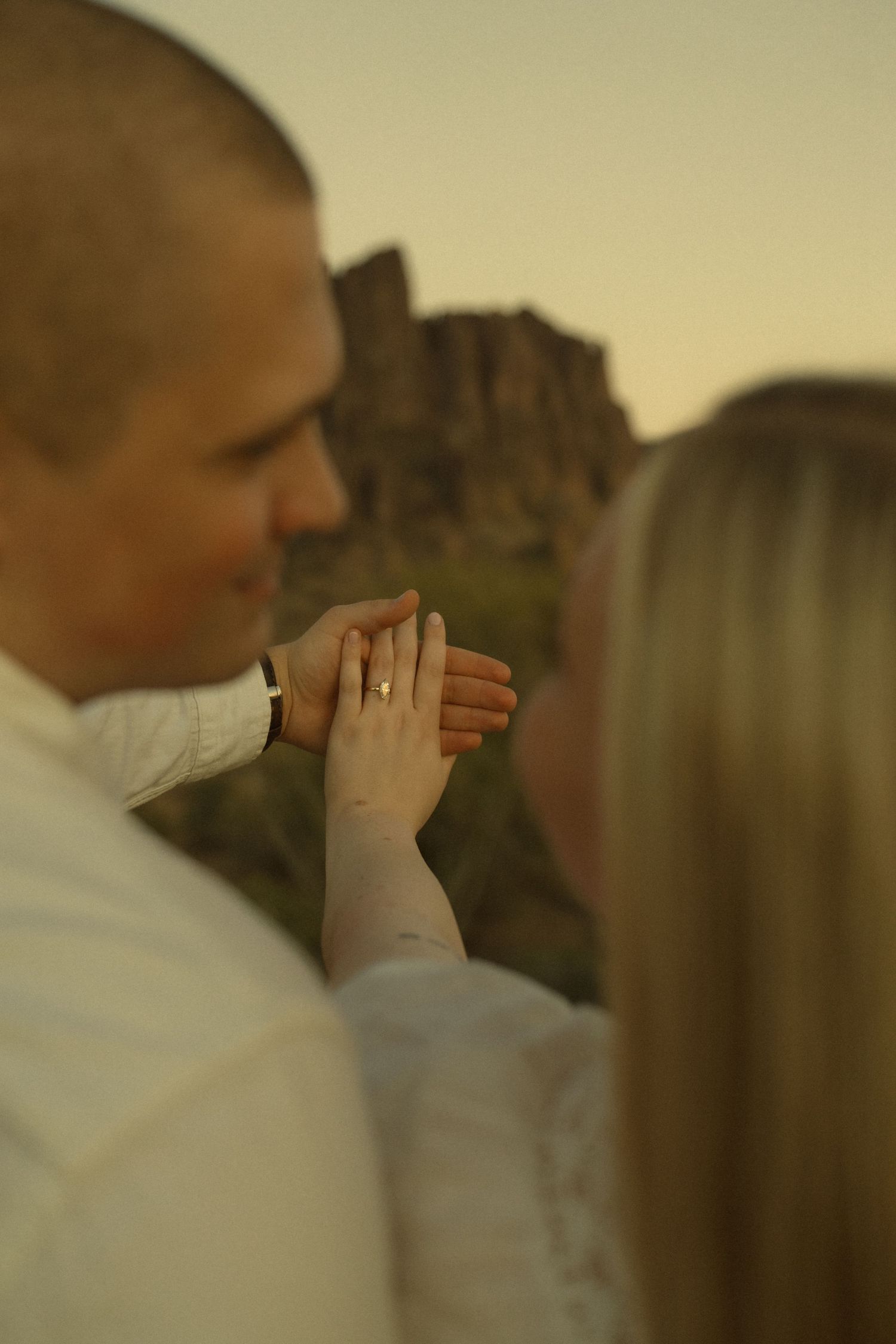 Couple holding hands showing engagement ring outdoors at sunset with rocky mountain landscape in background.
