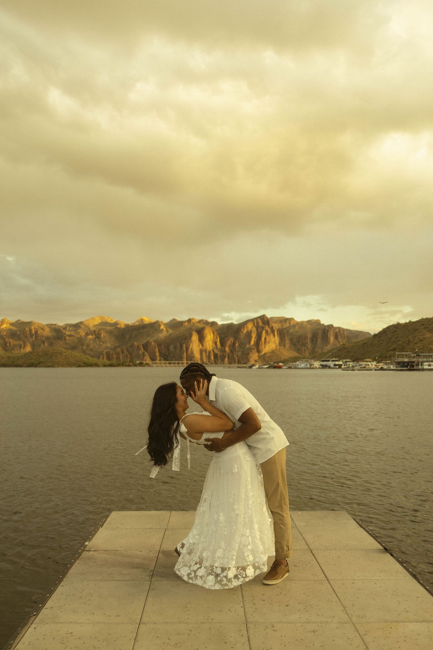 Couple sharing a romantic dip kiss on a lake pier with sunlit rocky mountains at dusk.
