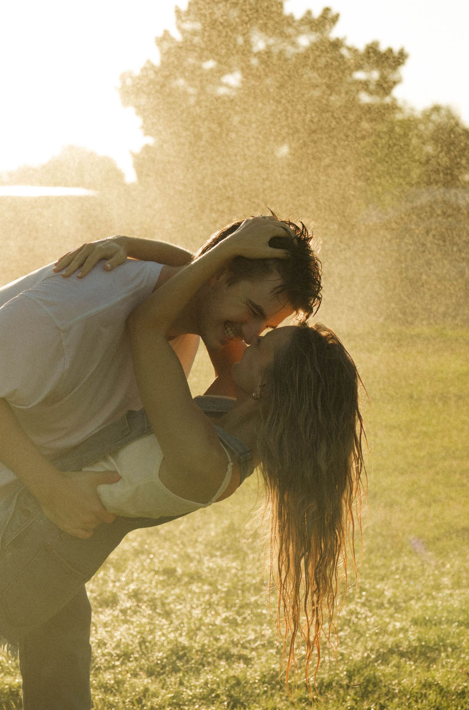 A couple embraces and nearly kisses while getting soaked by a sprinkler in a sunny green park.