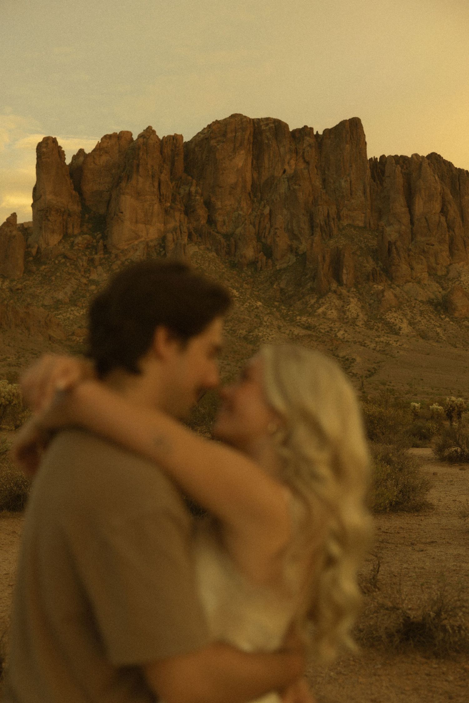 Couple sharing a romantic kiss in the desert with dramatic rocky mountain backdrop at golden hour.
