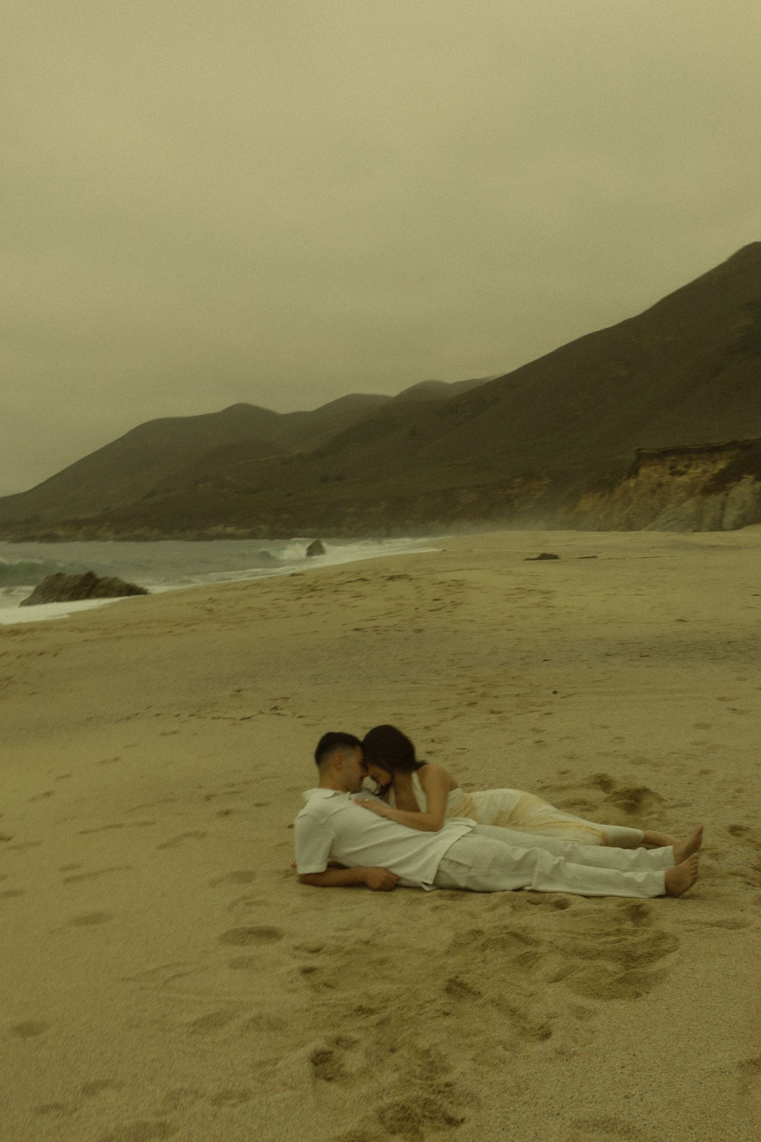 Couple lying together on sandy beach with dramatic coastal cliffs and moody overcast sky.