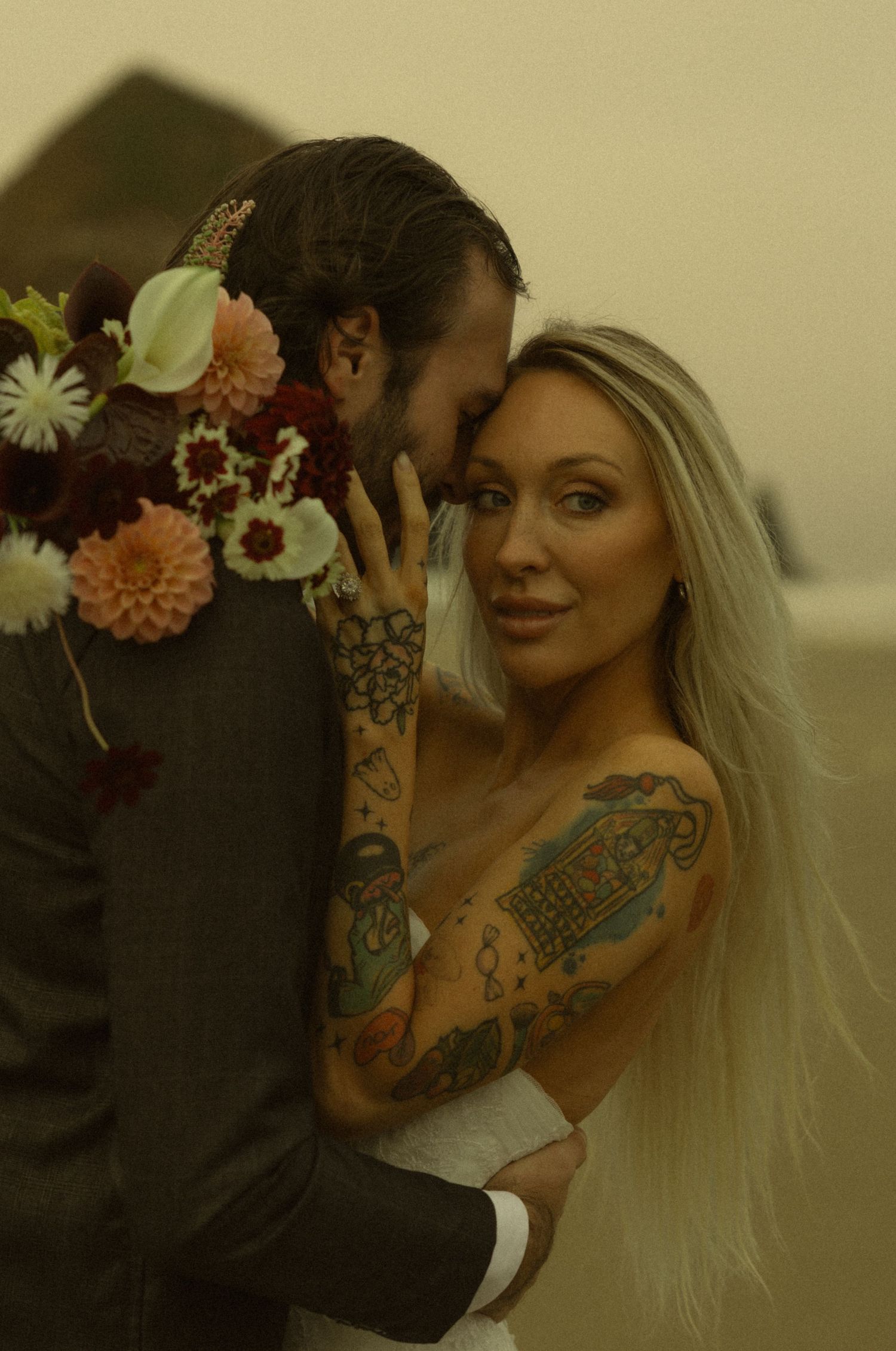 Tattooed bride in white dress embraces groom holding colorful floral bouquet during romantic beach wedding ceremony.