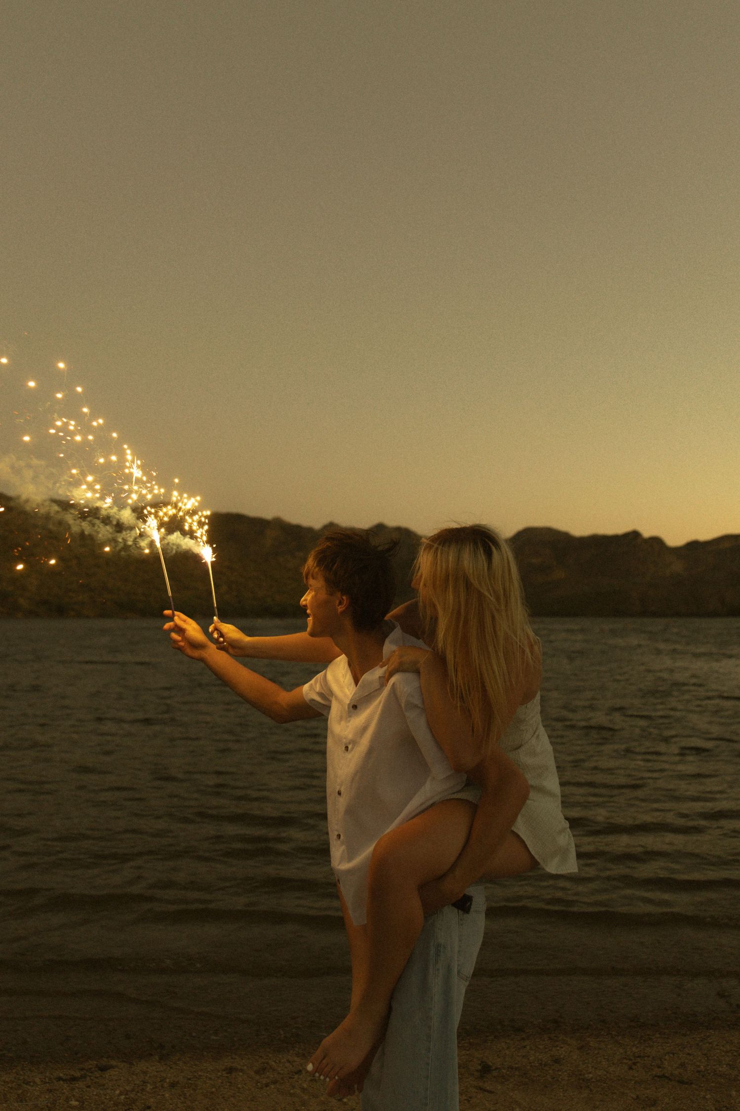 Couple holding sparklers on beach at sunset, man carrying woman on his back near lake with mountain backdrop.