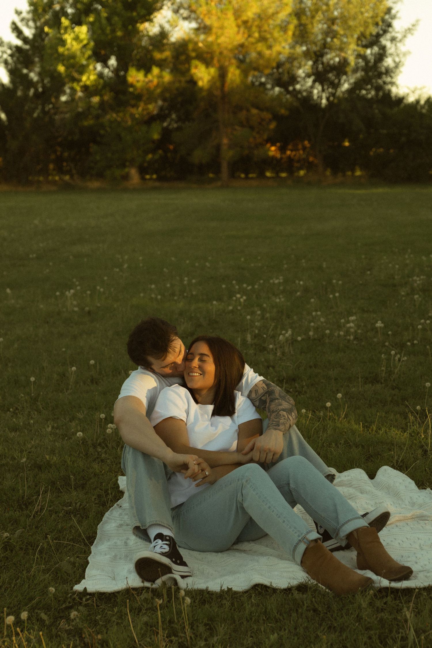Couple sitting on a white blanket in a sunny park, sharing a tender moment as one leans in for a kiss.