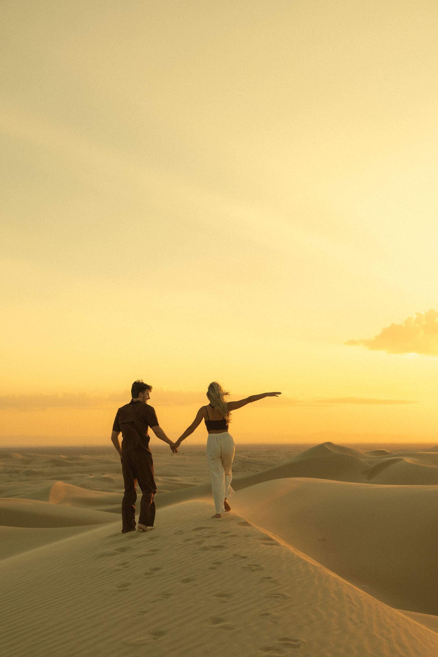 Couple holding hands on golden sand dunes at sunset in the desert, woman reaching out gracefully.