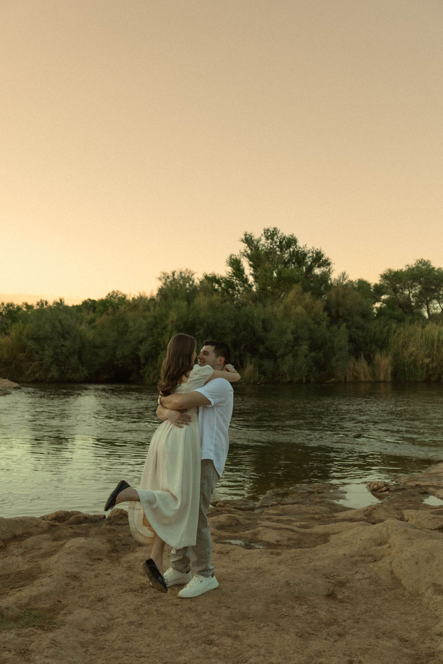 Man lifting woman in white dress by riverside at golden hour sunset.