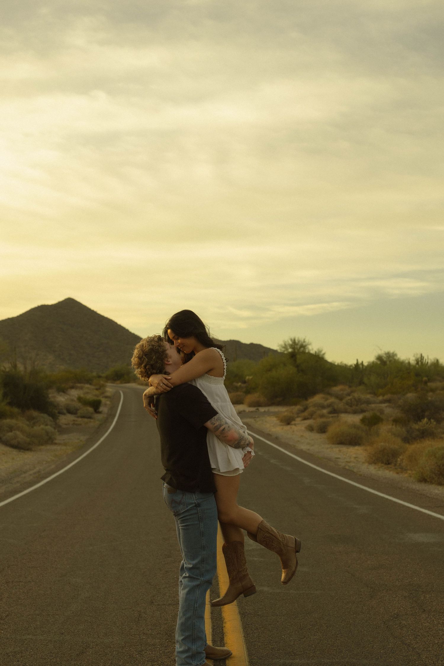 Man lifting woman in romantic embrace on empty desert highway at golden sunset with cacti landscape.