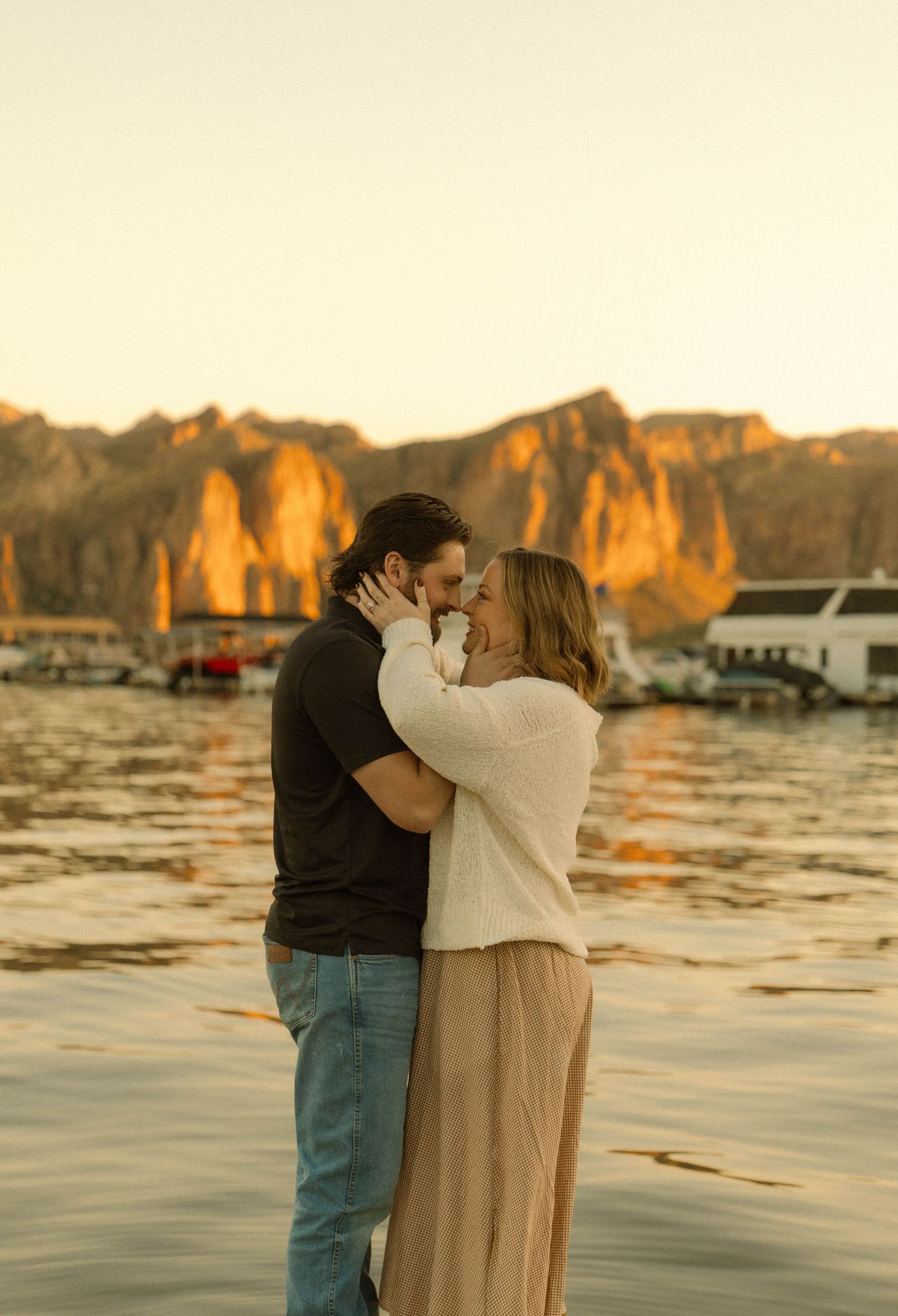 A romantic couple embraces at sunset by a lake with golden cliffs and mountains in the background.