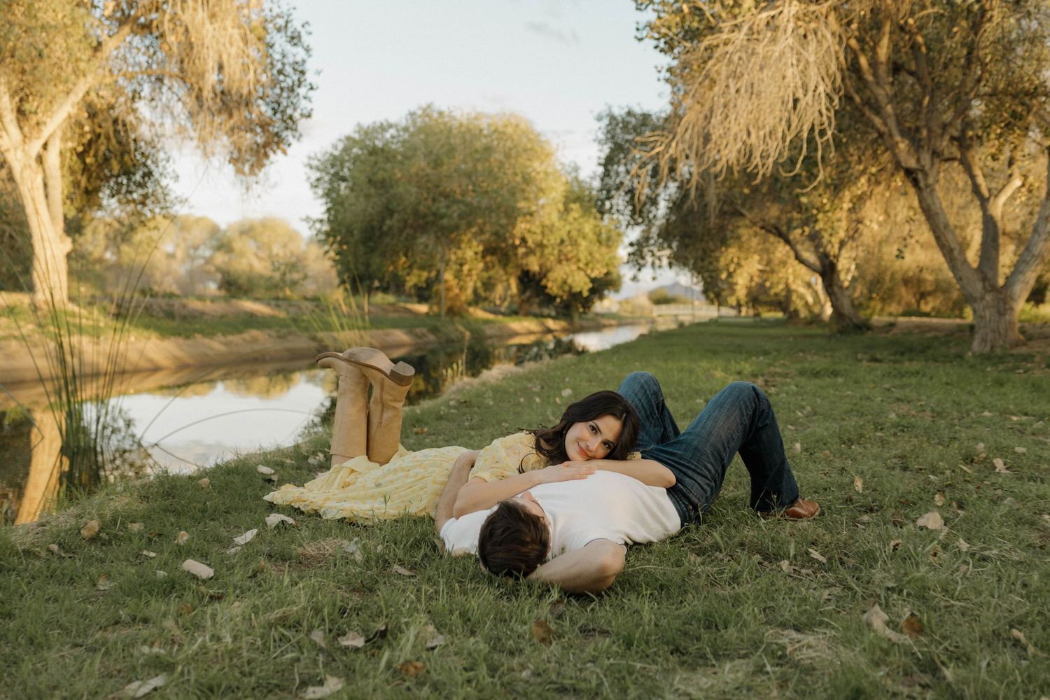 A couple relaxes together on grassy ground near a pond with trees creating a peaceful outdoor scene.