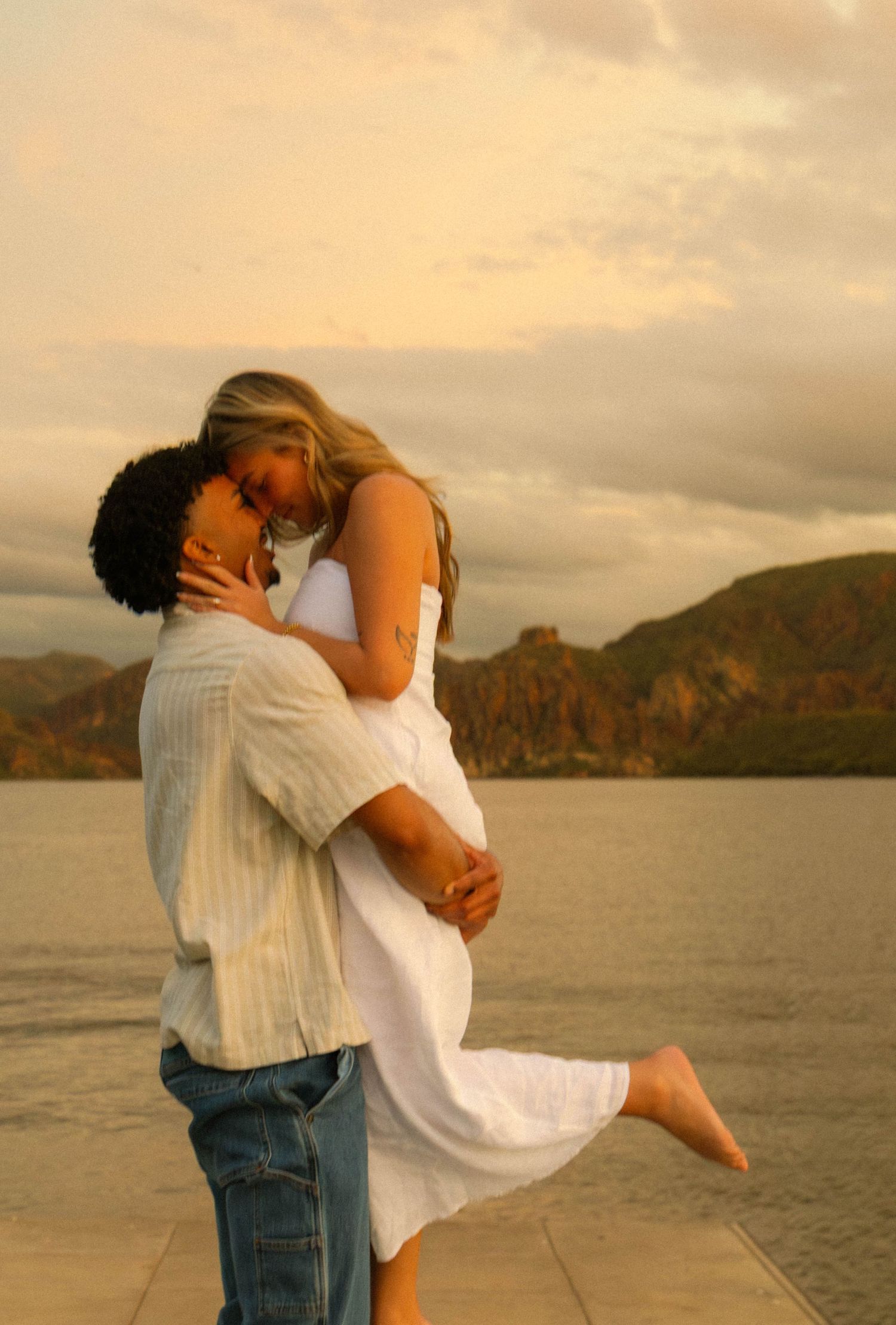 A romantic couple embraces at sunset by the ocean with mountains in the background, creating a silhouette effect.