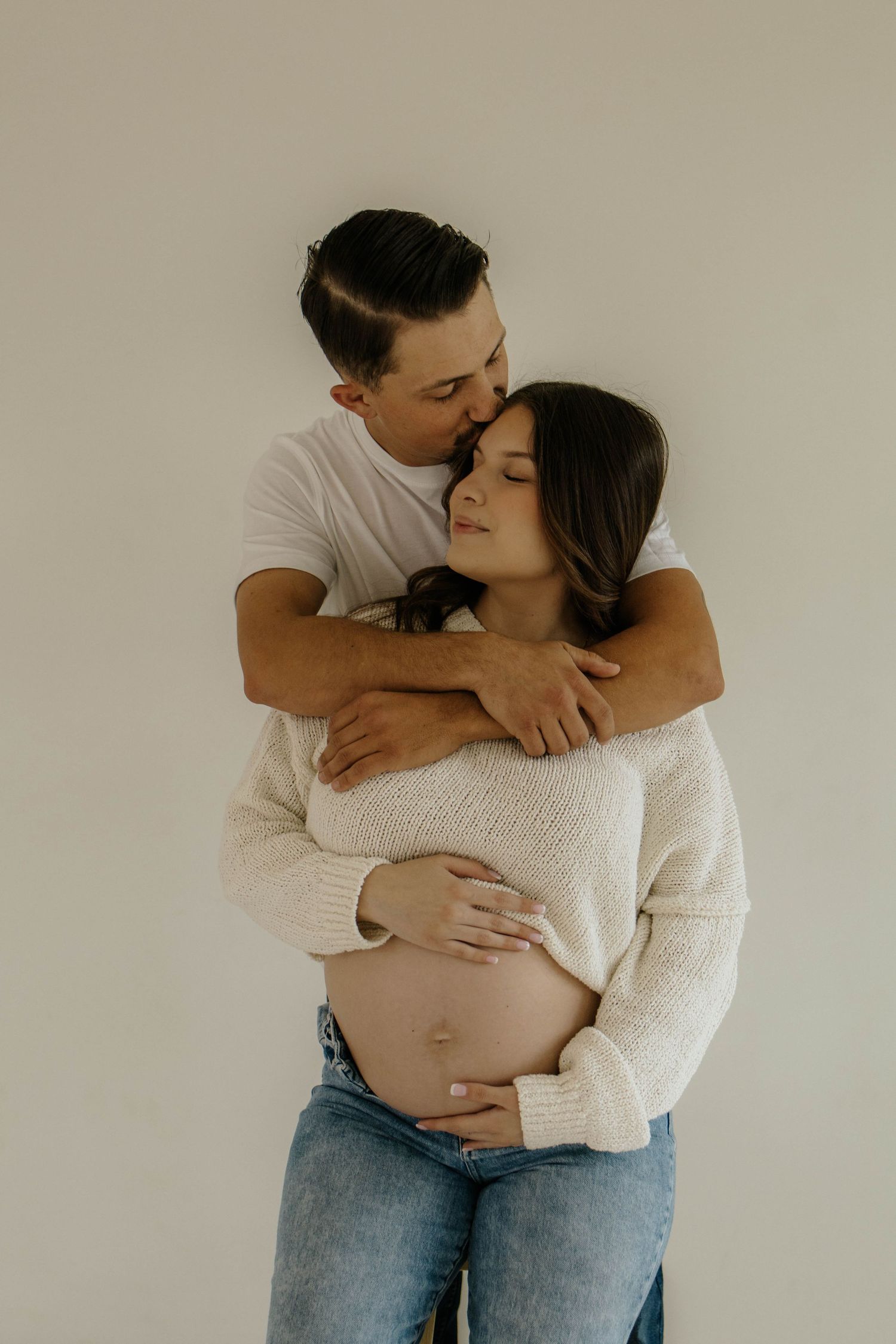 An intimate maternity photoshoot showing a couple in white and denim sharing loving moments.