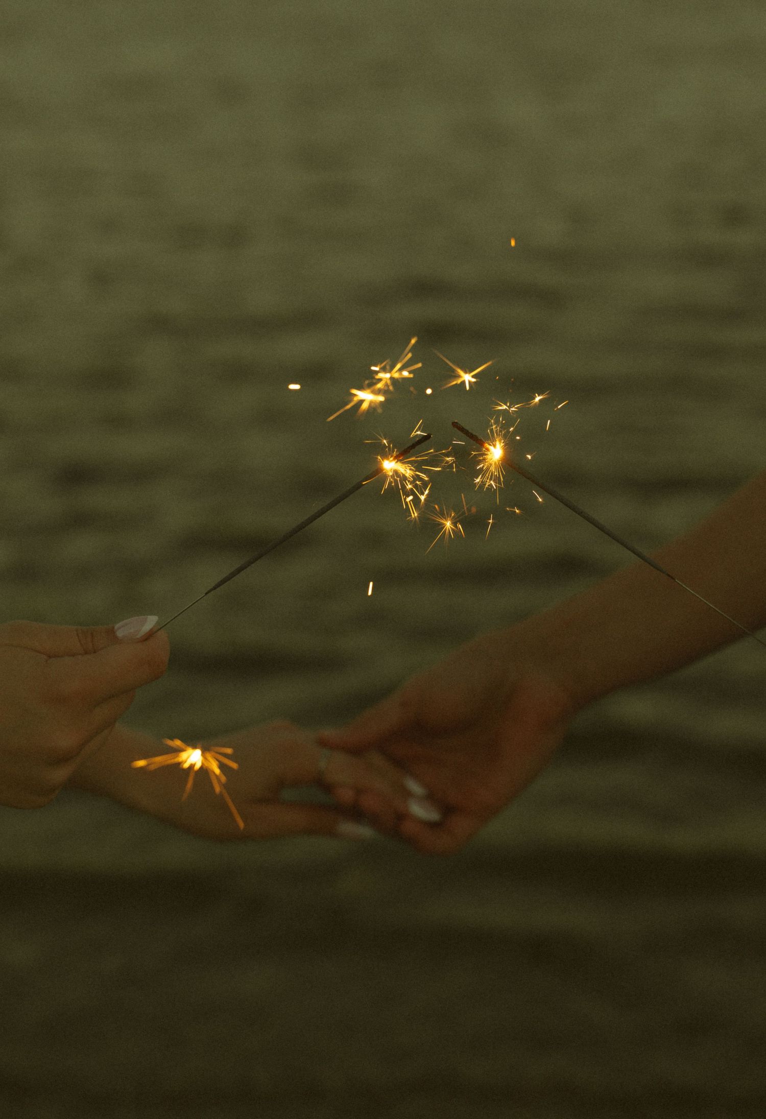 Two hands holding sparklers near water at dusk, with golden sparks flying between them in a romantic outdoor setting.