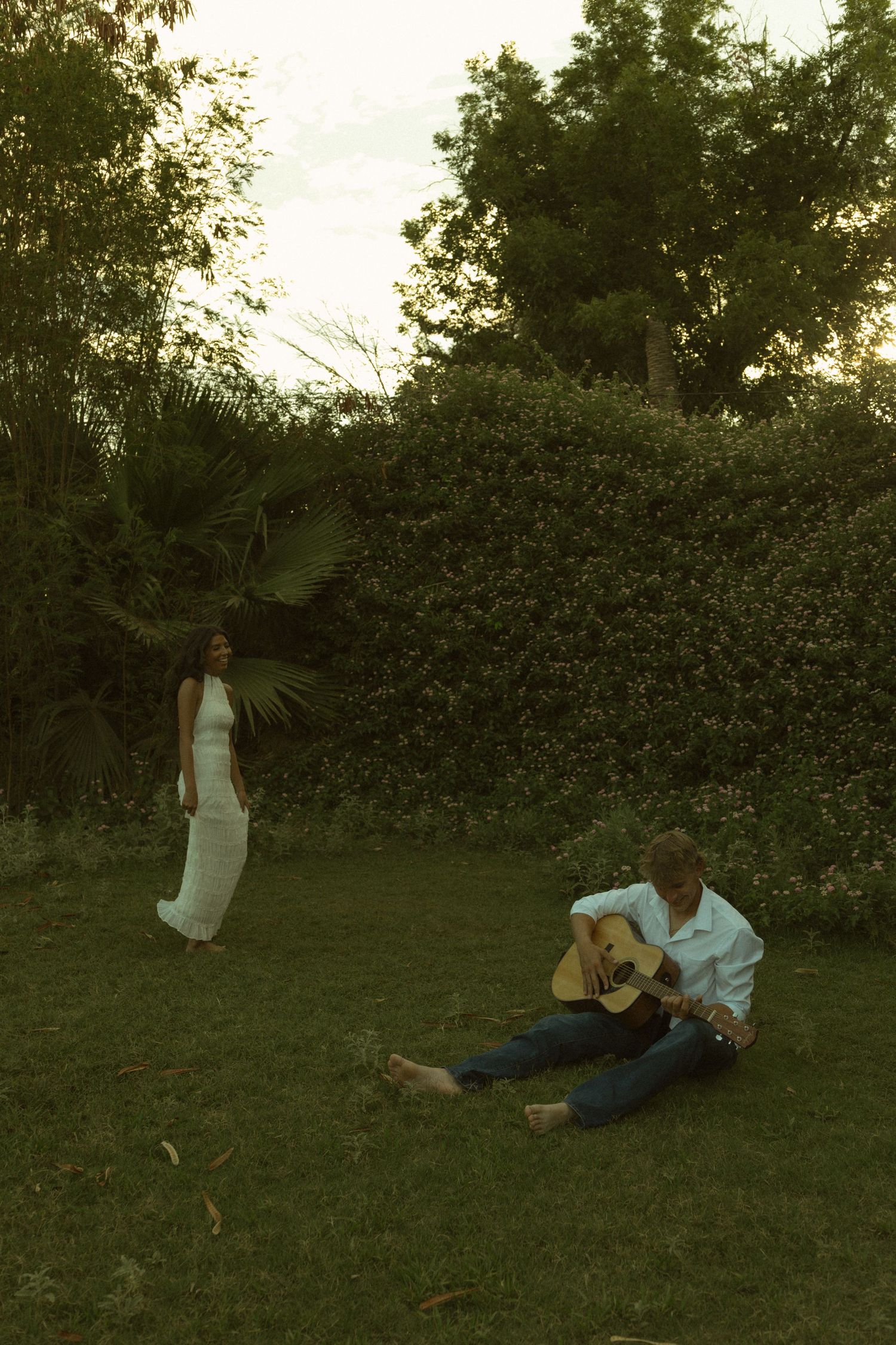 Man playing acoustic guitar on grass serenades woman in white dress standing in a lush green garden at dusk.