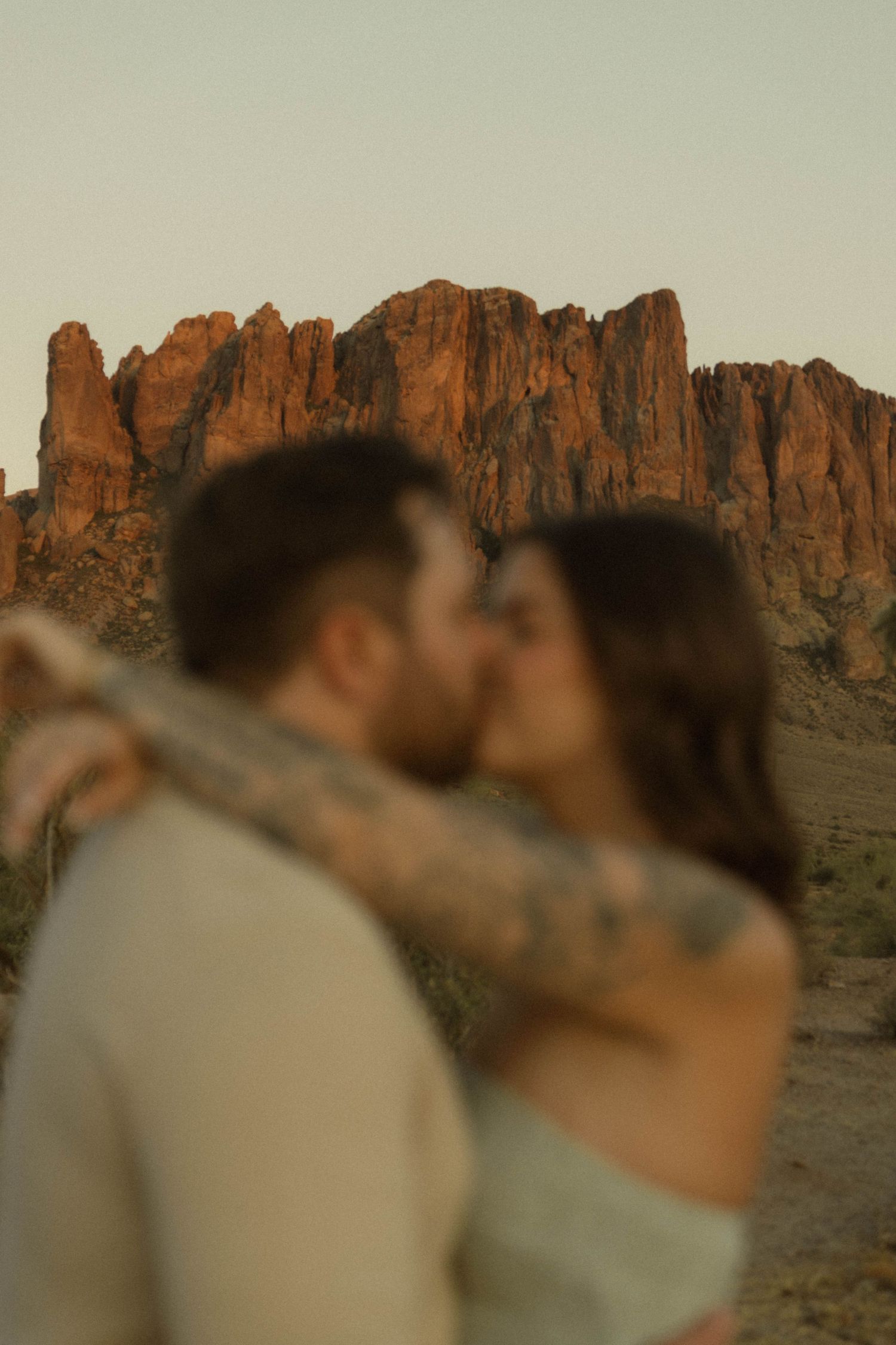 Couple sharing a romantic kiss with blurred tattoed arms and stunning red rock cliffs at golden hour.