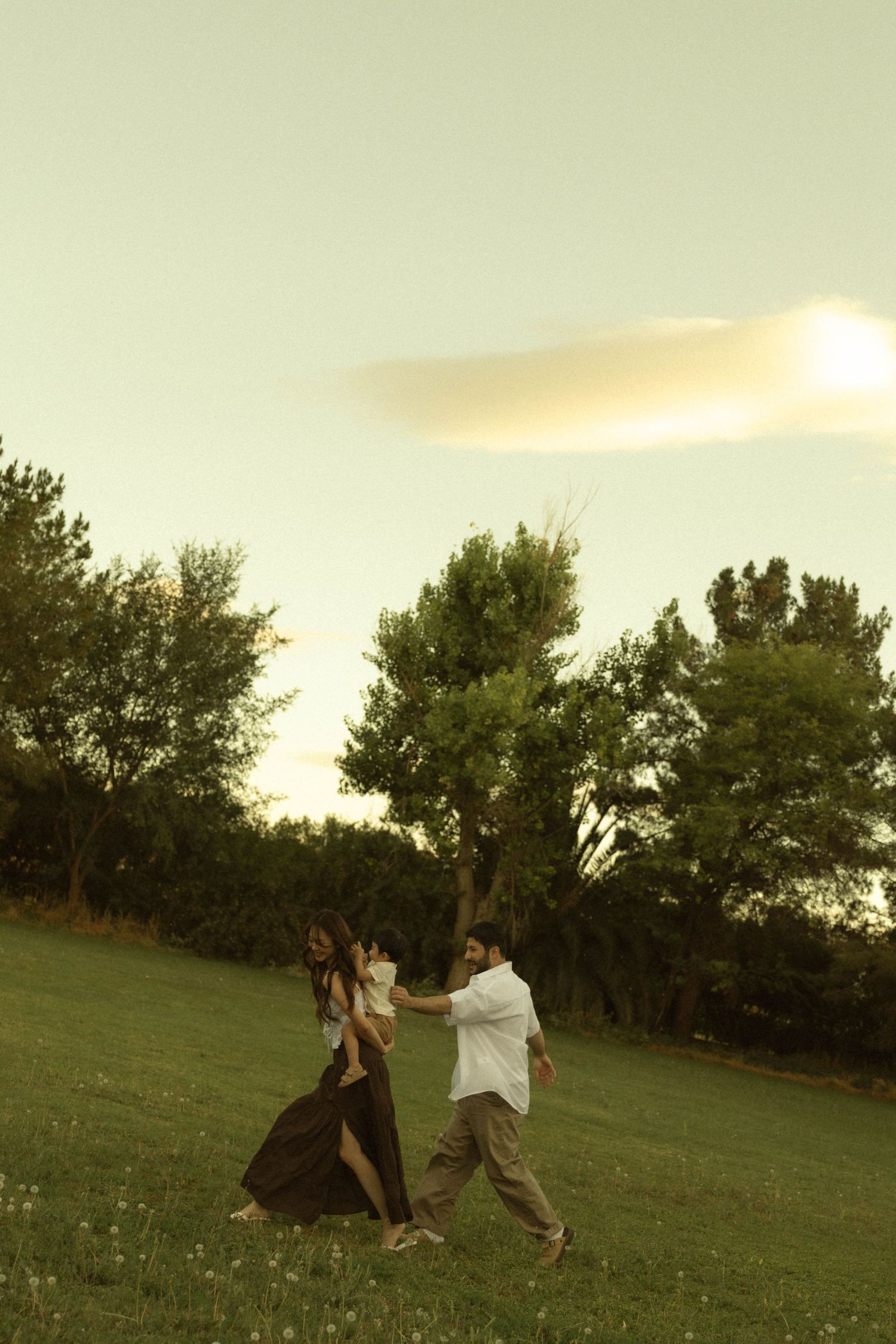 A couple walks together on a grassy field at golden hour, carrying their child amid scenic trees.