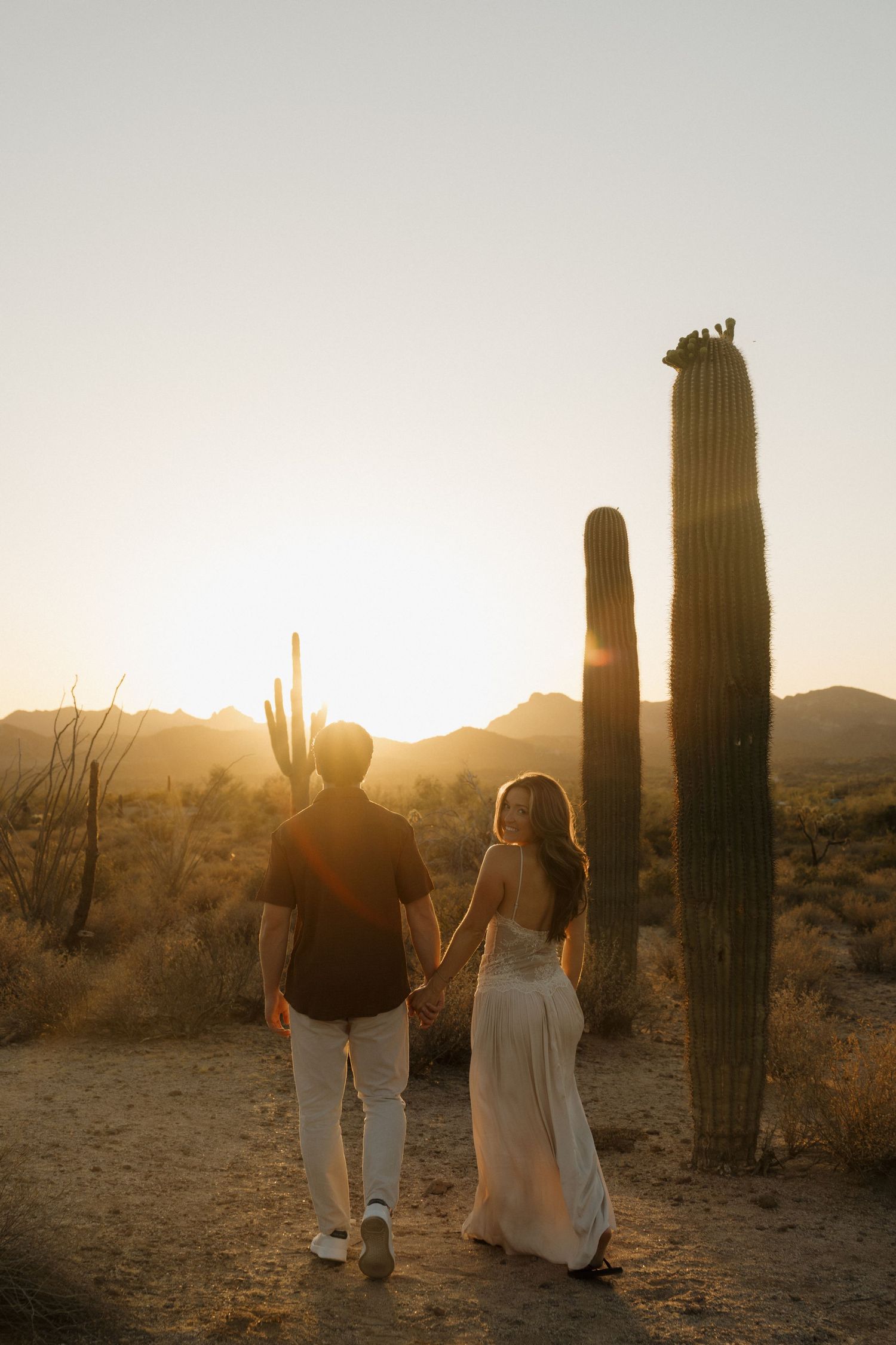 Couple holding hands walking toward the sunset among towering saguaro cacti in the Arizona desert landscape.