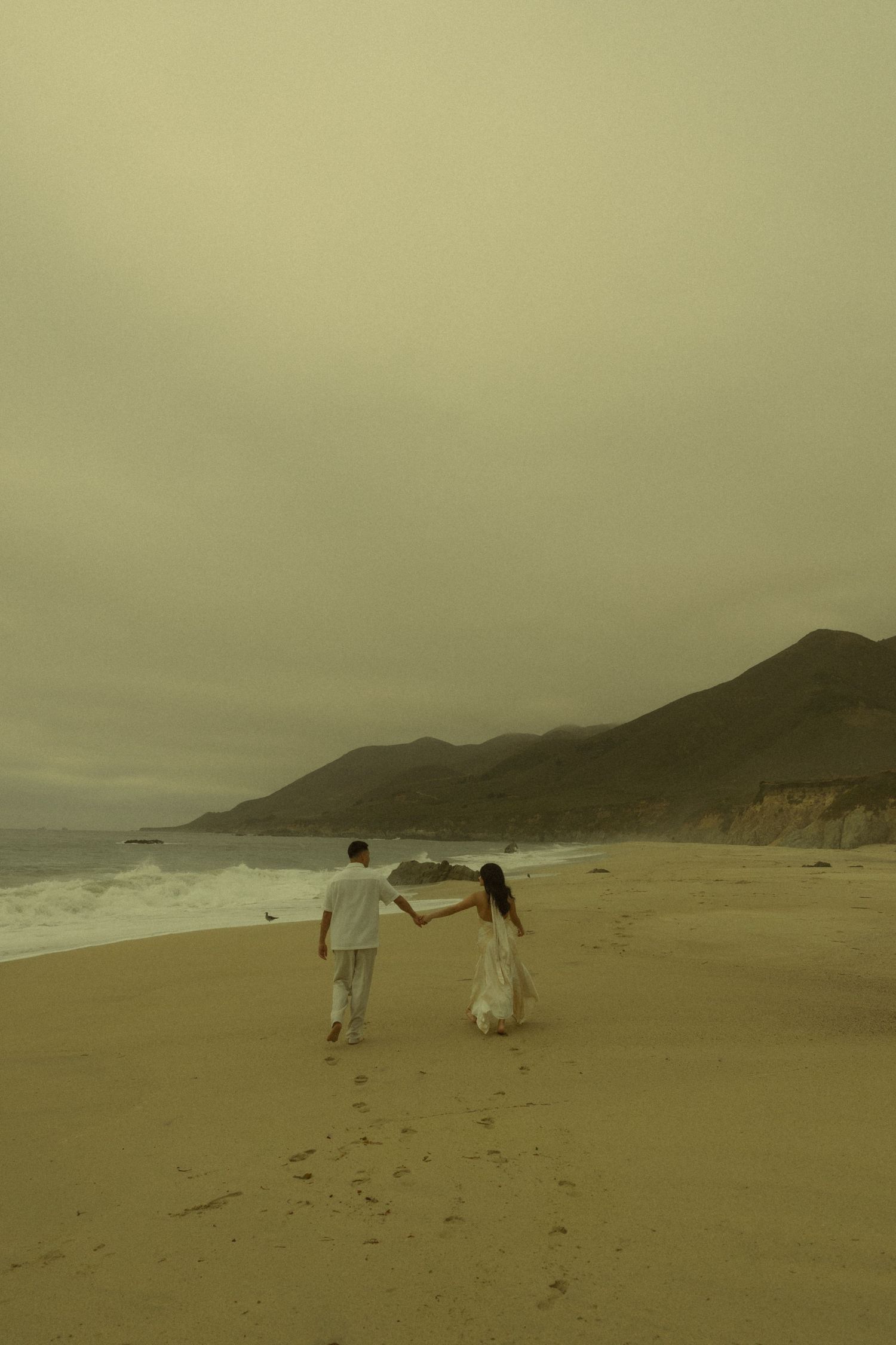 Couple in white holding hands walking on a sandy beach with ocean waves and misty mountains in the background.
