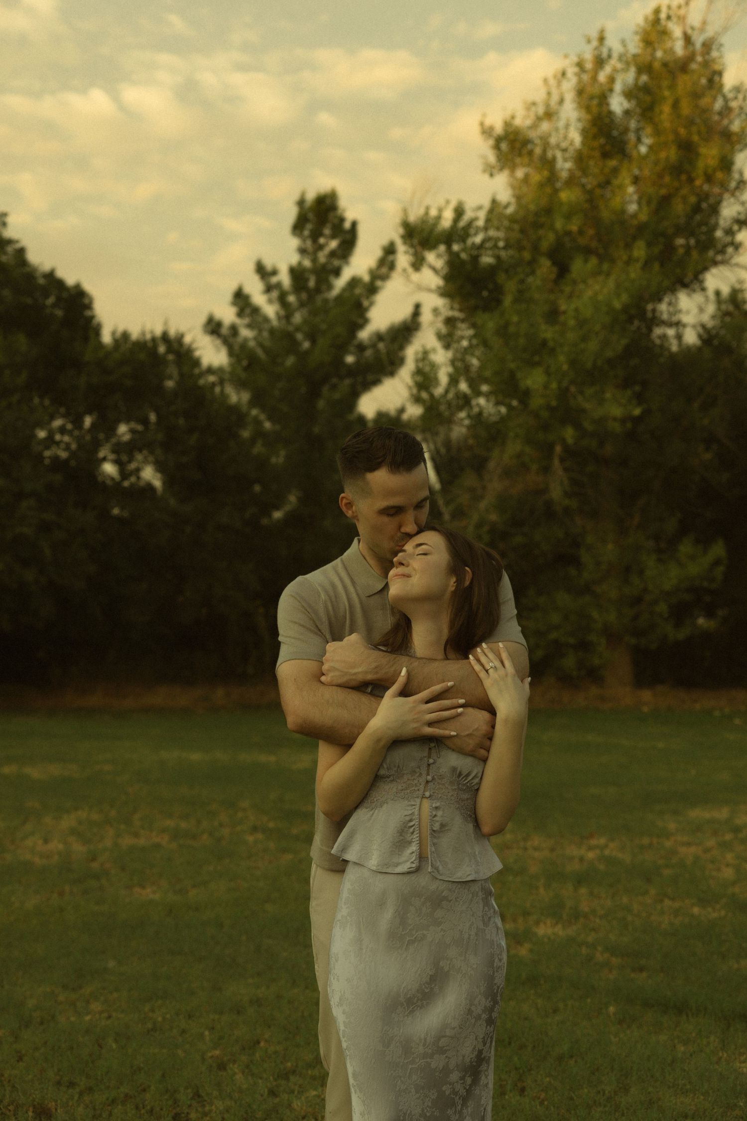 Couple embracing tenderly in a lush green park at golden hour, woman in white dress with eyes closed.