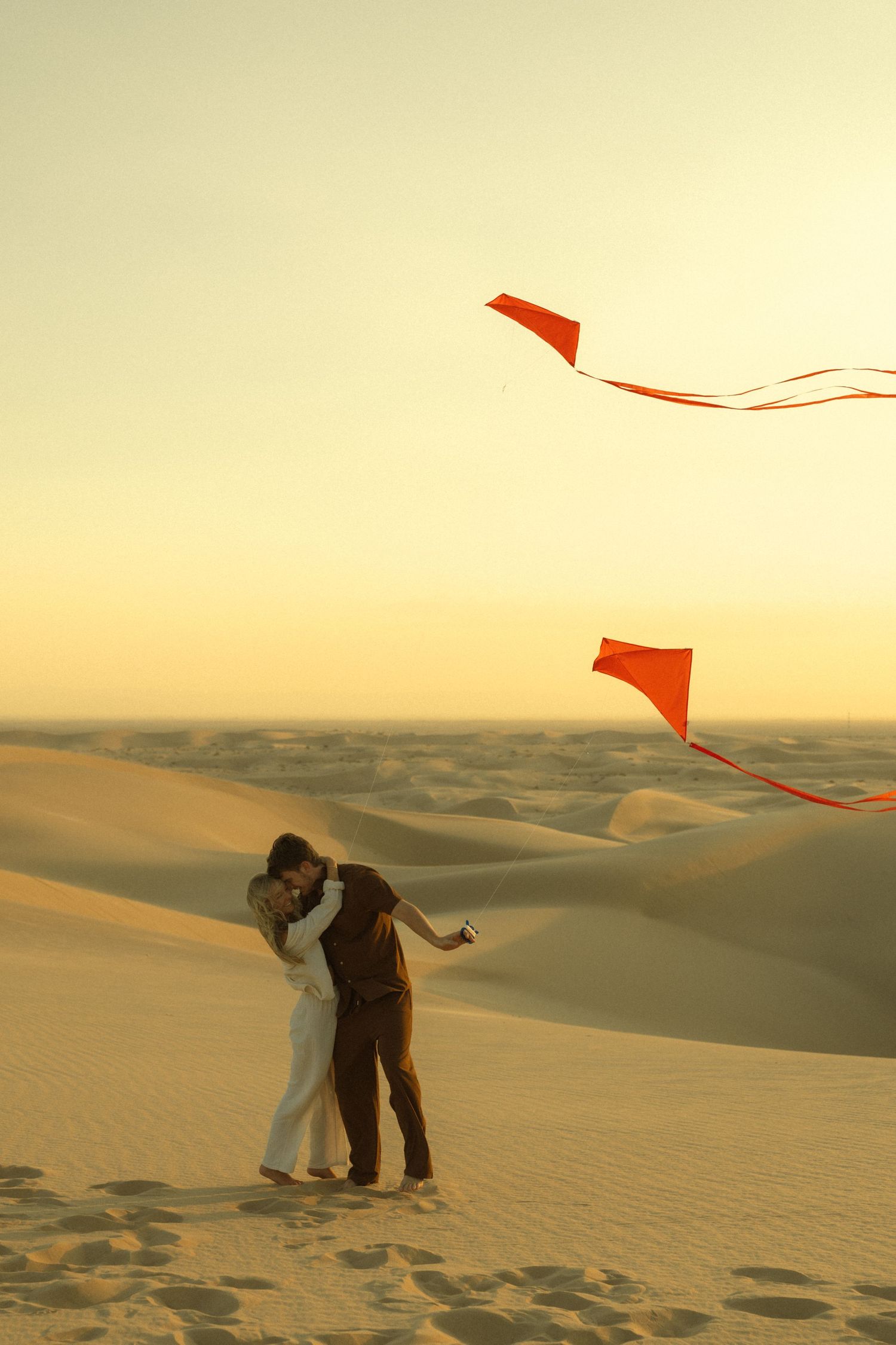Couple embracing on golden sand dunes at sunset with two red kites flying in the sky.
