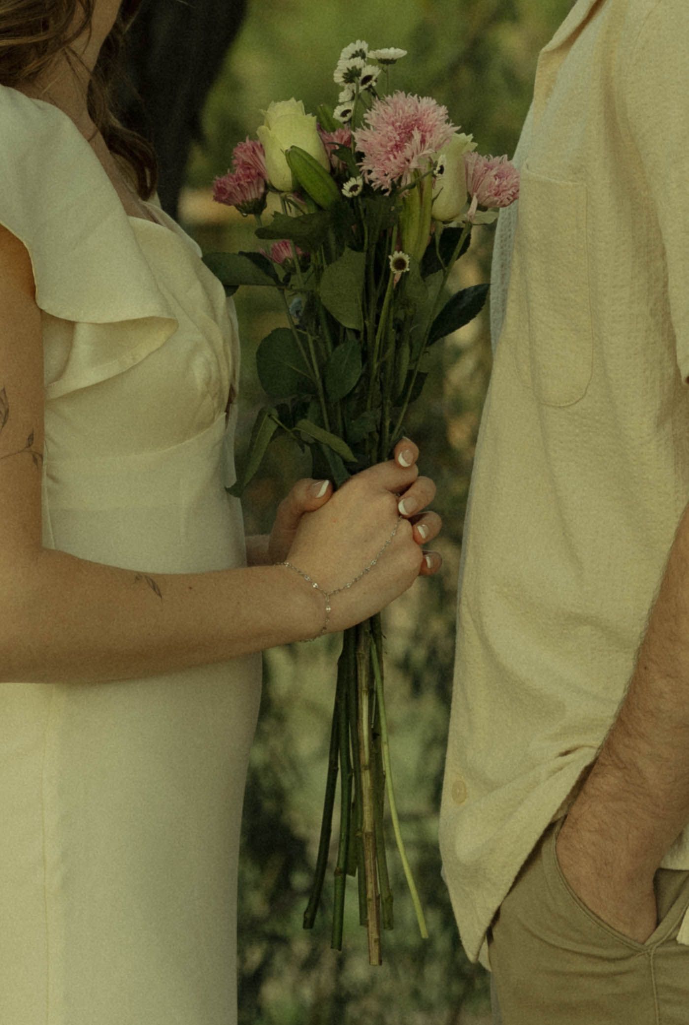Couple holding a bouquet of pink and white flowers together, wearing cream outfits, in a romantic outdoor setting.