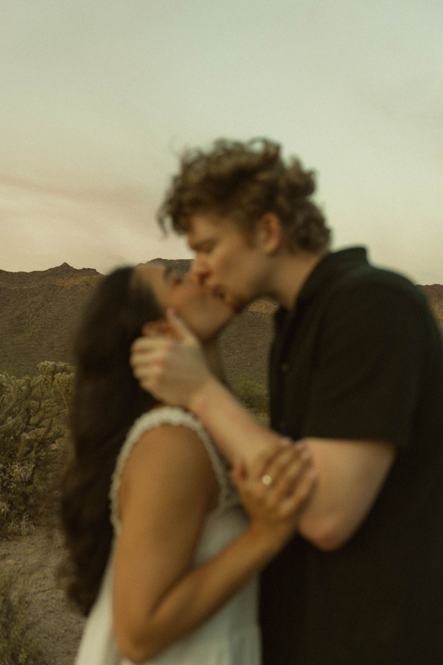 Blurred artistic photo of couple kissing at dusk with desert landscape and mountains in the background.