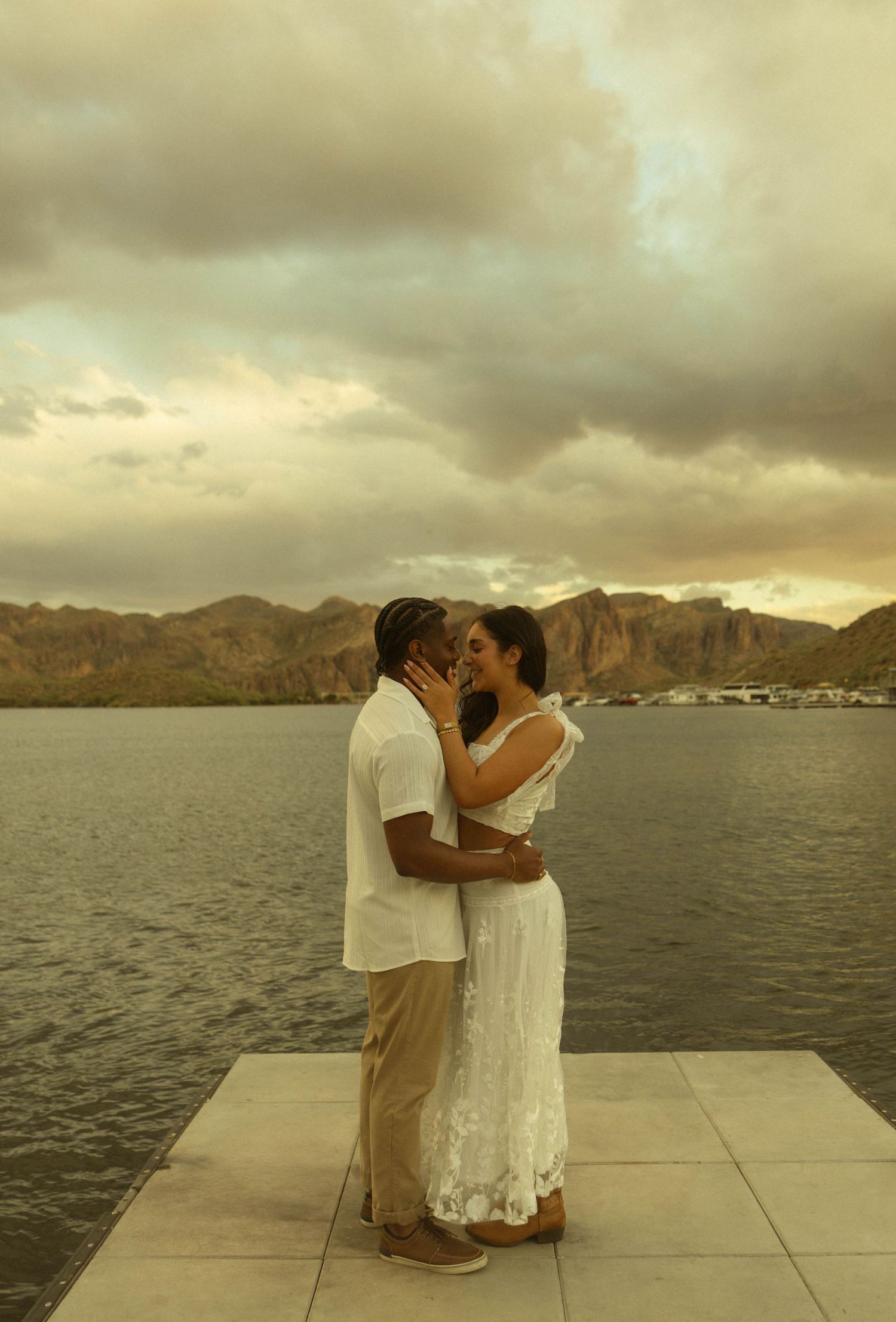 Couple sharing a tender kiss on a waterfront pier with dramatic cloudy sky and mountains behind.