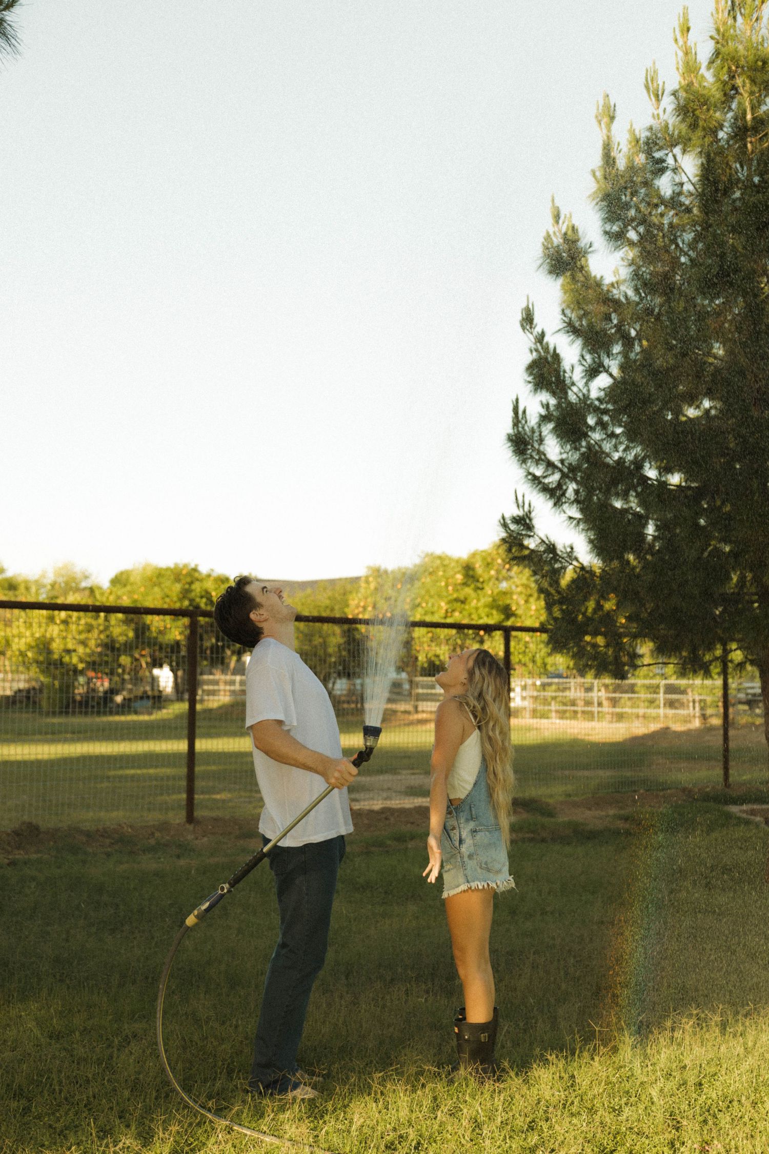 A couple plays with a garden hose in a sunny backyard near a wooden fence and tall evergreen tree.