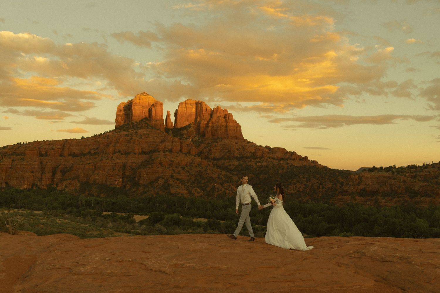 Bride and groom holding hands at sunset near Cathedral Rock in Sedona, Arizona, with golden clouds and red rock formations.