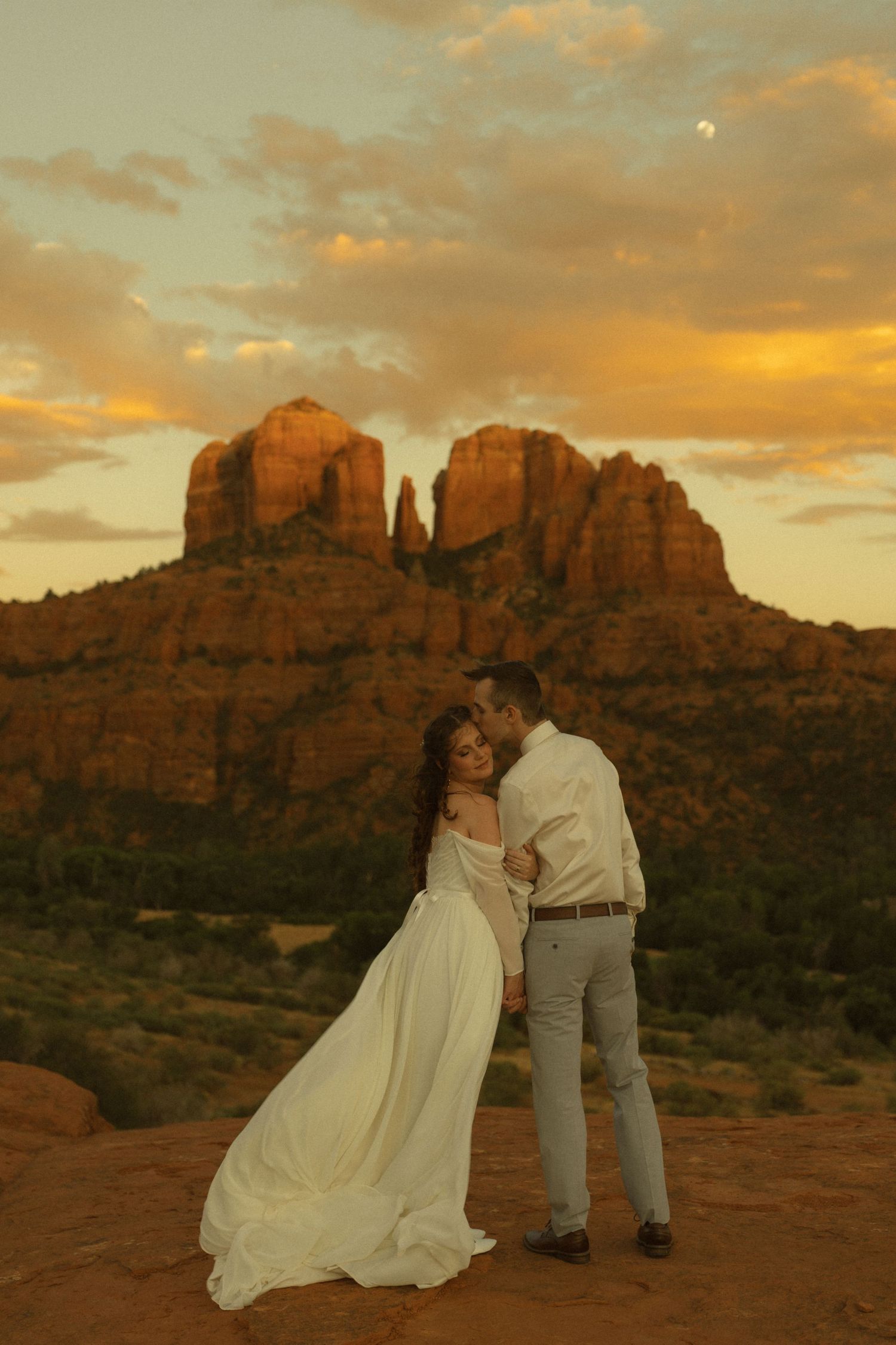 Couple embracing at sunset with Cathedral Rock in Sedona, Arizona, bride in flowing white gown, golden sky backdrop.